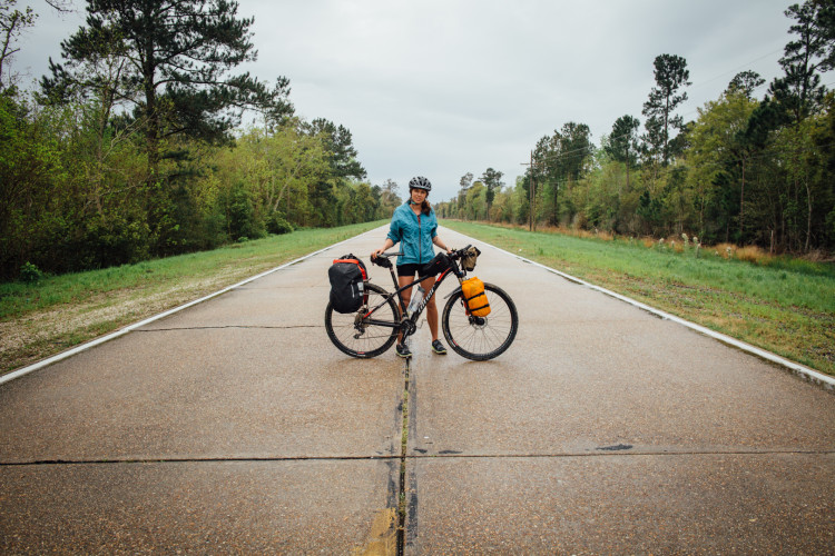 A person standing next to a bicycle on a quiet, empty road surrounded by trees. The individual is wearing a helmet and a blue jacket, holding the bike upright with bags attached for travel. The scene is set under a cloudy sky, indicating overcast weather.