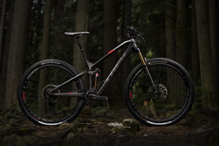 A sleek black mountain bike with red accents is positioned on rocky terrain, surrounded by tall trees in a dimly lit forest. The bike features a lightweight frame, high-quality tires, and a front suspension fork, highlighting its design for off-road performance.