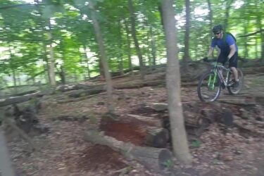 A mountain biker riding through a wooded trail, navigating over fallen logs and rough terrain, surrounded by lush green trees. Novi Tree Farm (Lakeshore Park) mountain bike trail.