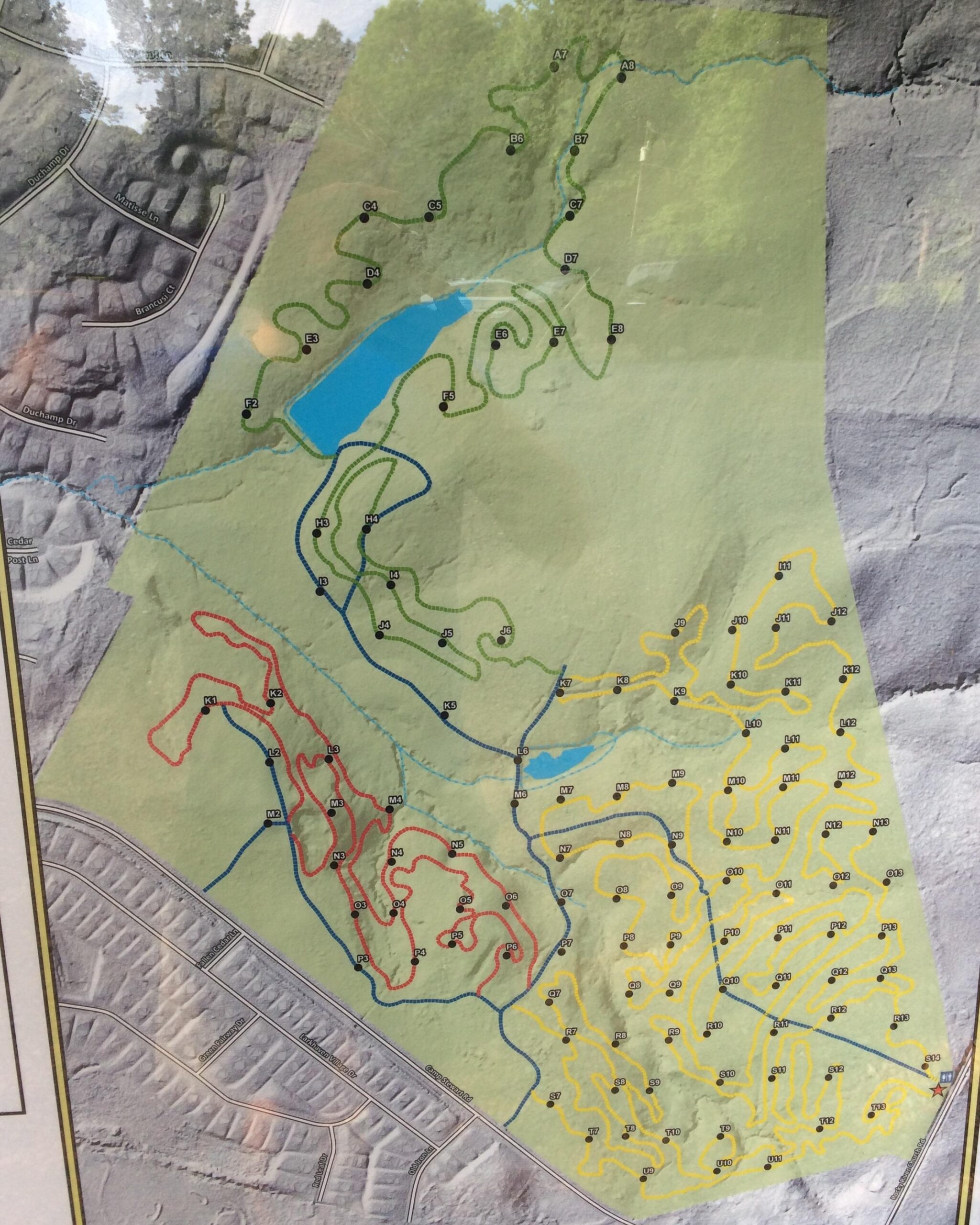 Map displaying hiking or biking trails, featuring colored paths in green, blue, red, and yellow. The map includes a small pond and various marked trail points across the terrain, with surrounding residential areas noted in the bottom left corner. Sherman Branch Park Mtb Trail mountain bike trail.