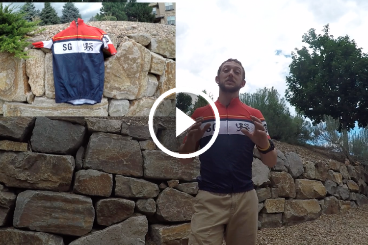 A man in a cycling jersey is standing in front of a stone wall, gesturing as he talks. To the left, there is an image of a cycling jersey hanging on the wall. The background features trees and a partly cloudy sky.