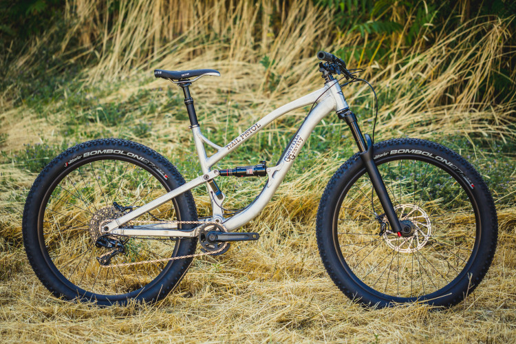 A silver mountain bike with wide tires stands on a bed of straw, showcasing its sleek frame and suspension system. The bike features a prominent seat and handlebars, and is set against a backdrop of green foliage.
