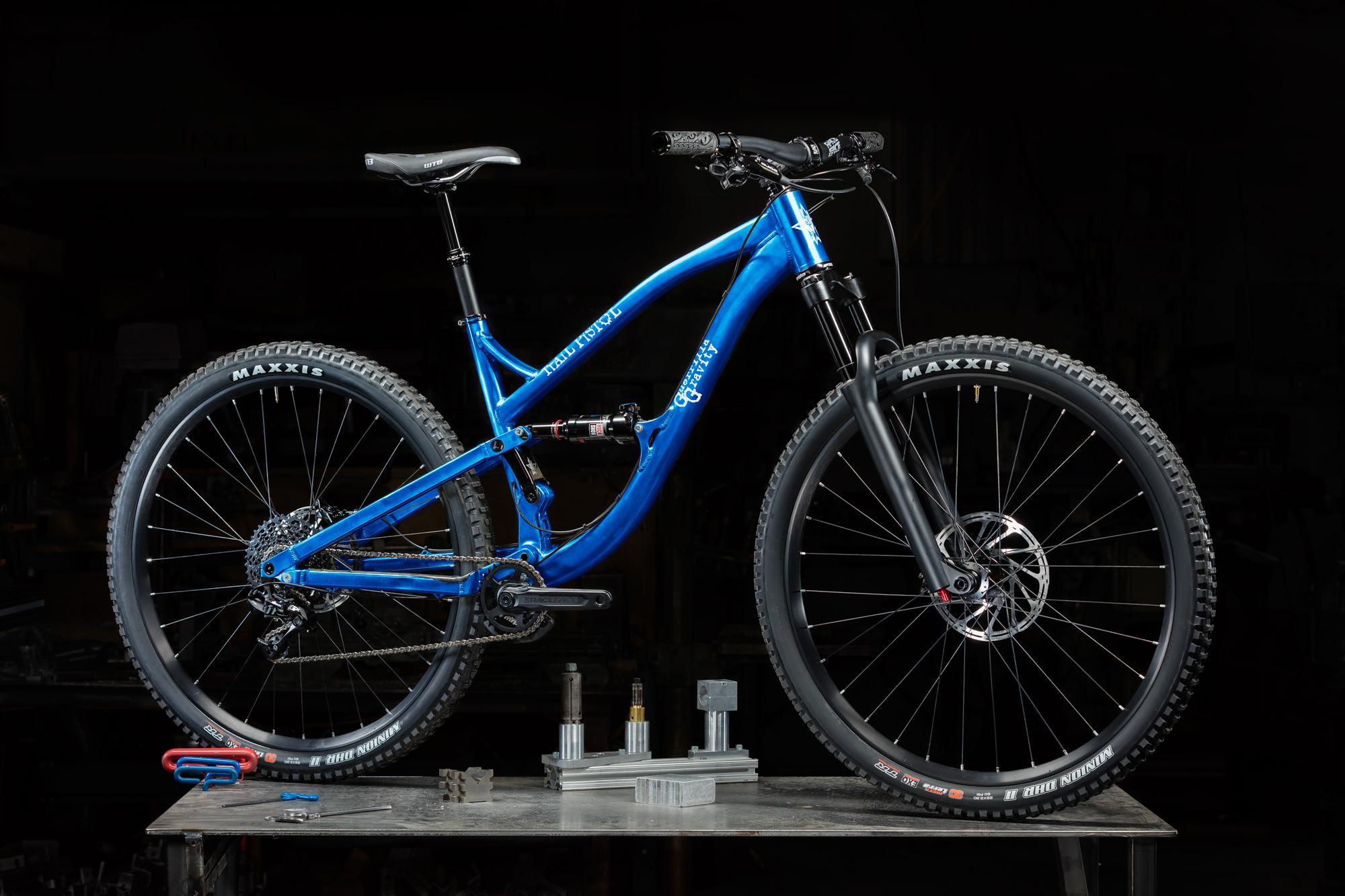 Guerrilla Gravity Trail Pistol: A blue mountain bike displayed on a workbench, featuring large knobby tires, a modern frame design, and various mechanical components in the background. The bike is positioned against a dark backdrop, emphasizing its sleek appearance and details.