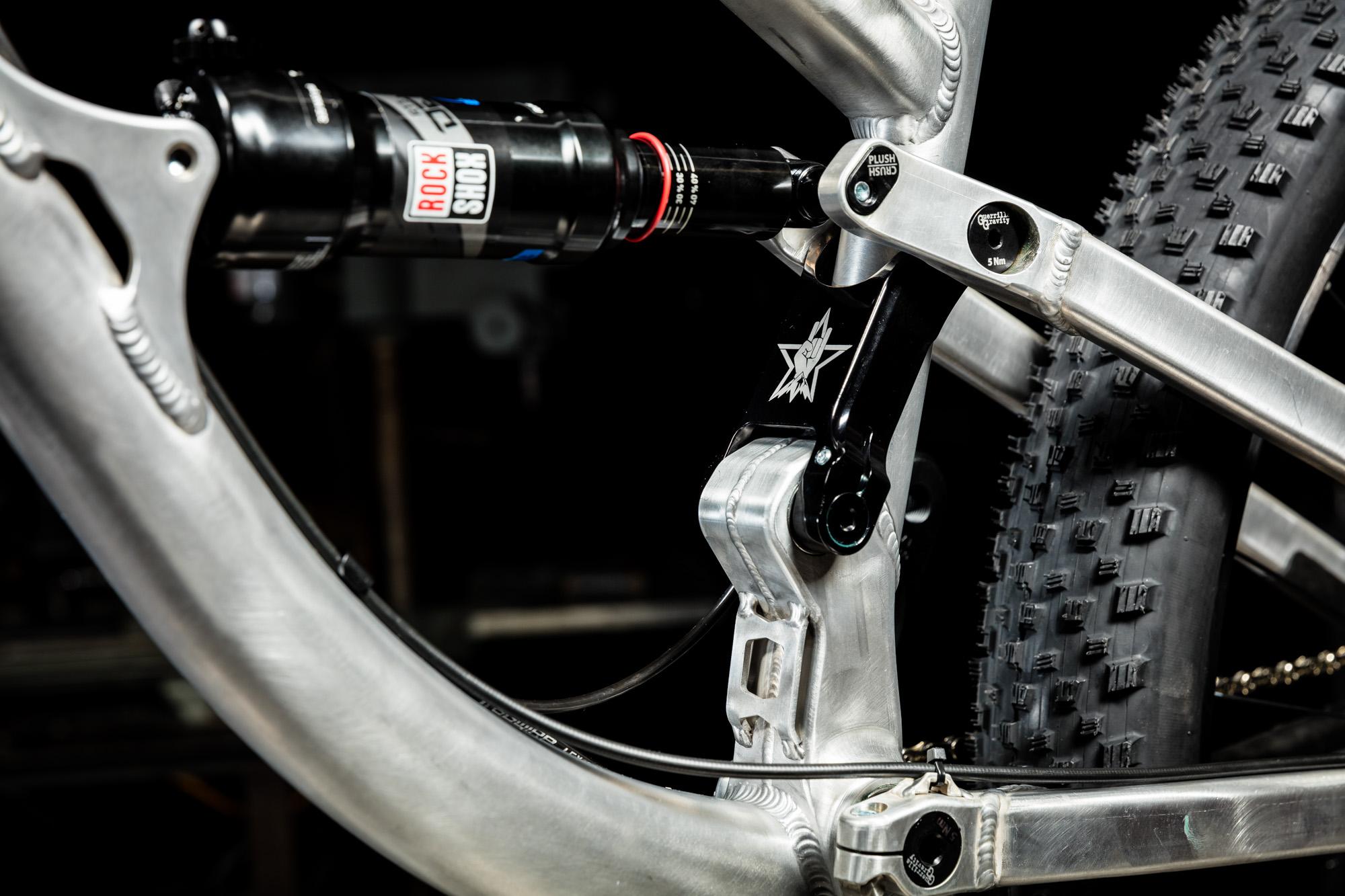 Guerrilla Gravity Trail Pistol: Close-up of a mountain bike's rear suspension system, featuring a shock absorber and metallic frame components. The image highlights detailed craftsmanship with an aluminum finish and visible tire tread in the background, set against a dark backdrop.