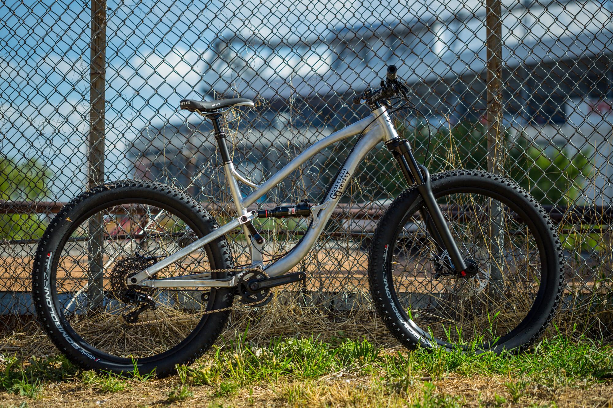 Guerrilla Gravity Trail Pistol: A silver mountain bike leaning against a chain-link fence, with large black tires and a visible suspension system. The background includes a blurred structure and patches of grass and dry plants.