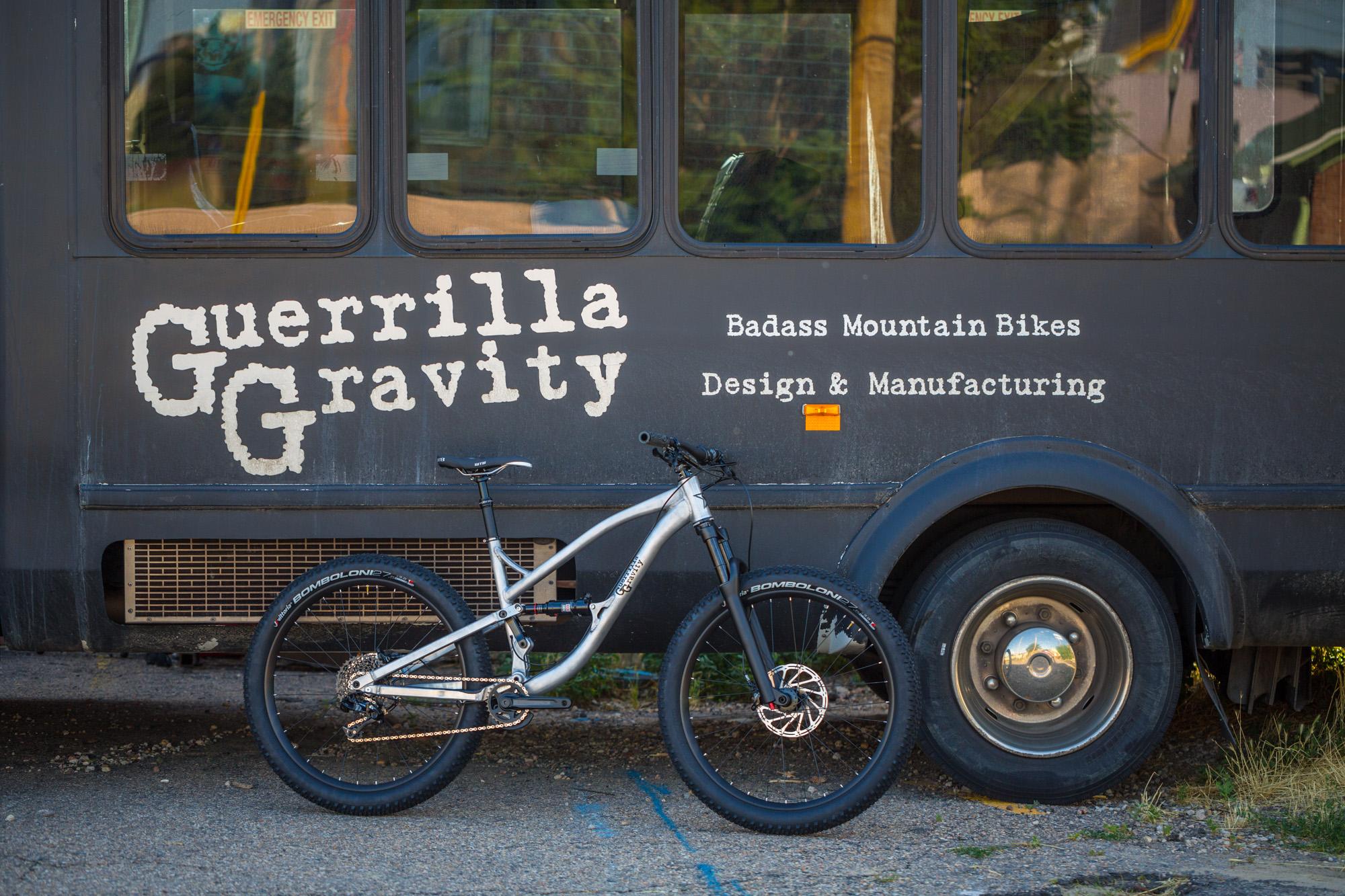 Guerrilla Gravity Trail Pistol: A silver mountain bike leans against a black bus featuring the text "Guerrilla Gravity" along with "Badass Mountain Bikes" and "Design & Manufacturing." The bus has an industrial look and is parked on a gravel surface.