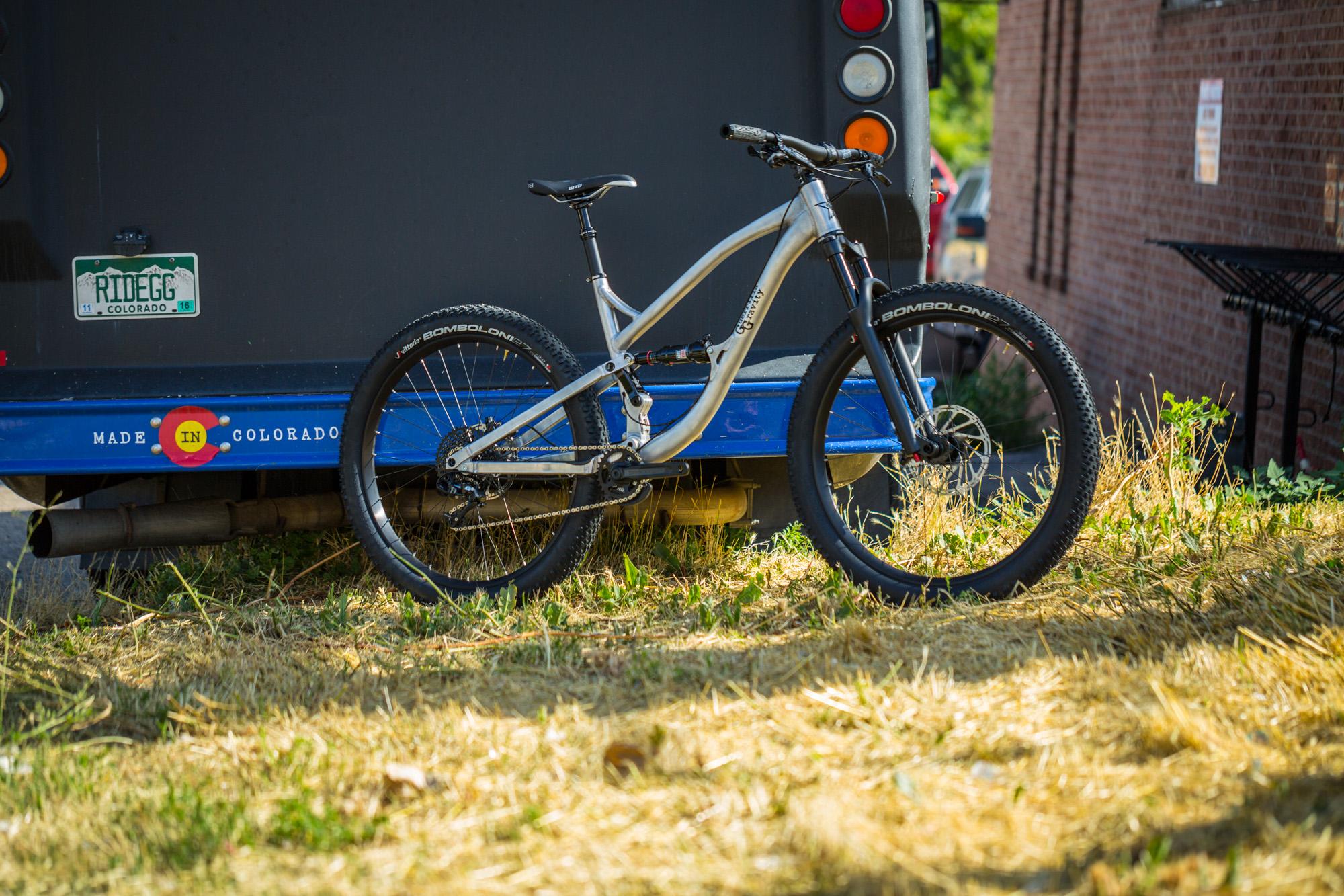 Guerrilla Gravity Trail Pistol: A mountain bike with a silver frame and thick tires is leaning against a blue truck. The scene is set in a grassy area, with a portion of a brick wall visible in the background. The truck has Colorado license plates and a sign that reads "MADE IN COLORADO." Sunlight casts a warm glow on the bike and surroundings.