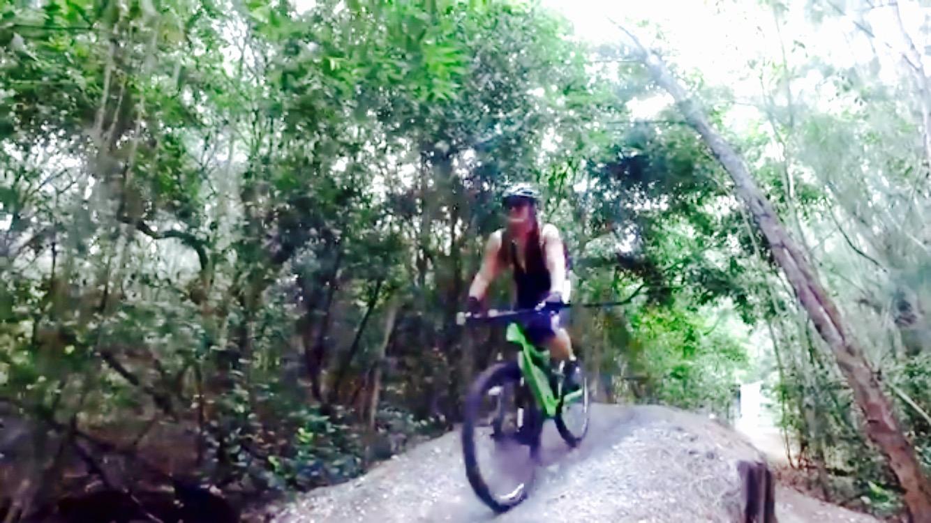 A person biking along a gravel trail in a lush green forest, surrounded by dense trees and foliage. The cyclist is wearing a helmet and riding a green mountain bike. Markham Park mountain bike trail.
