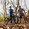 Giant Giant Trance 27.5 2: Two individuals stand next to their mountain bikes on a forest trail, surrounded by trees and fallen leaves. One person is wearing a blue shirt and the other is in a gray shirt. The setting appears to be a sunny day in nature.