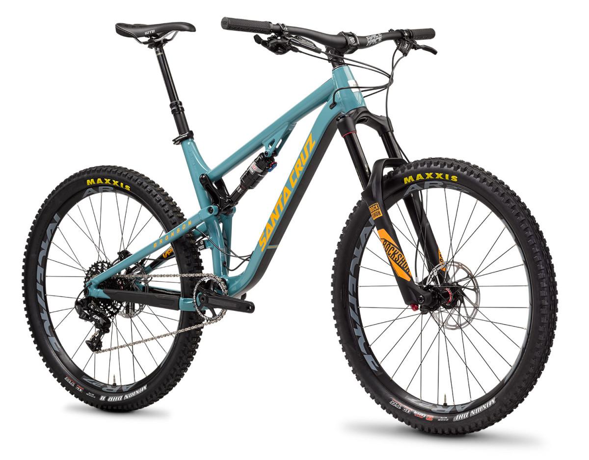 Santa Cruz Bronson Aluminum: A teal mountain bike with yellow accents, featuring a full-suspension frame, wide tires with Maxxis branding, and a prominent front fork. The bike has a sleek design, geared for trail riding, with detailed components and a modern aesthetic.