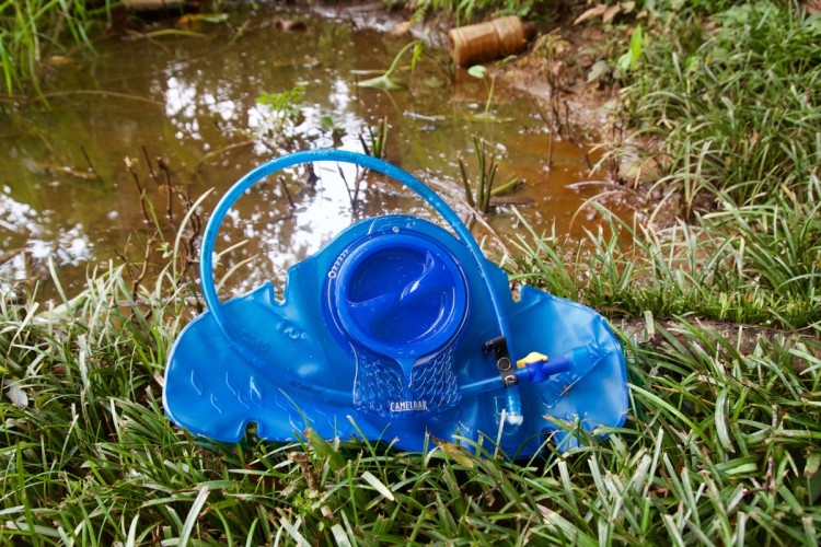 A blue hydration reservoir placed on green grass near a small, muddy body of water, with a drinking tube attached.