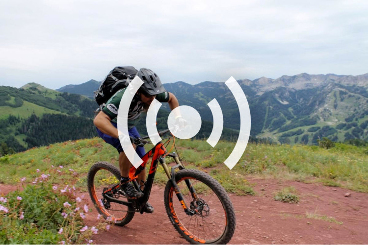 A mountain biker navigating a dirt trail through a scenic mountainous landscape, with lush greenery and distant hills in the background. The rider wears a helmet and a backpack, displaying an active outdoor lifestyle.