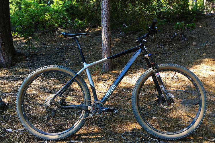 A black and silver mountain bike is positioned on a wooded forest floor, surrounded by trees and greenery. The bike features thick, knobby tires suitable for off-road trails and has visible components like a hydraulic fork and a rear cassette. The ground is covered with pine needles and scattered twigs, indicating a natural outdoor setting.