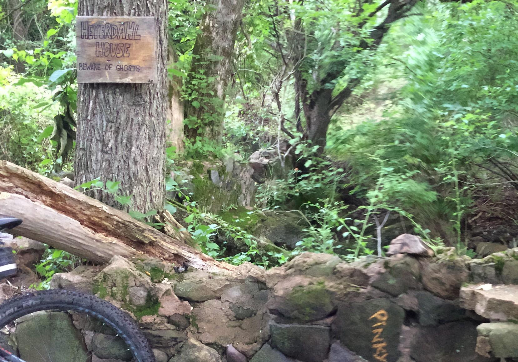 A wooden sign reading "HEYERDAHL HOUSE" with the phrase "BEWARE OF GHOSTS" hangs on a tree. The surroundings are lush and green, featuring a mix of trees and dense foliage. Stone remnants of a wall are visible in the foreground, and a bicycle's wheel is partially seen in the lower left corner. Richmond Avenue and Forest Hill road mountain bike trail.