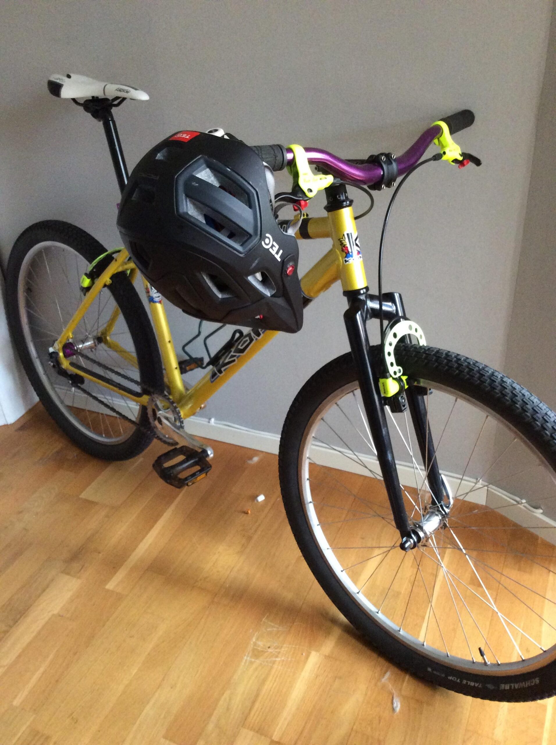 Kona Nunu: A yellow mountain bike with black tires and a purple handlebar, resting against a wall. A black helmet is positioned on the handlebars. The bike features a prominent fork and visible pedal. The background has wooden flooring and a gray wall.
