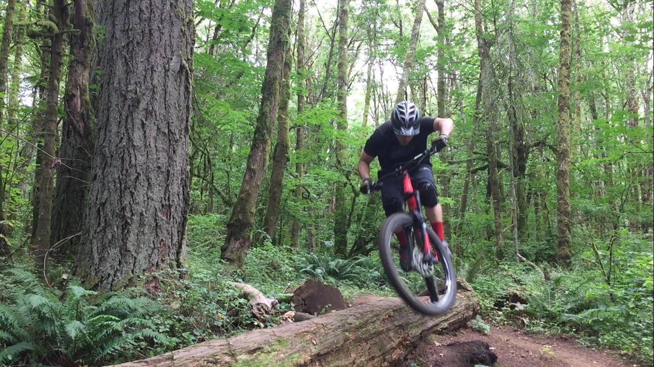 A mountain biker maneuvering over a fallen log in a lush green forest, surrounded by tall trees and ferns. The cyclist wears a helmet and sports gear, captured mid-jump with the bike's front wheel lifted off the ground. Round Lake mountain bike trail.