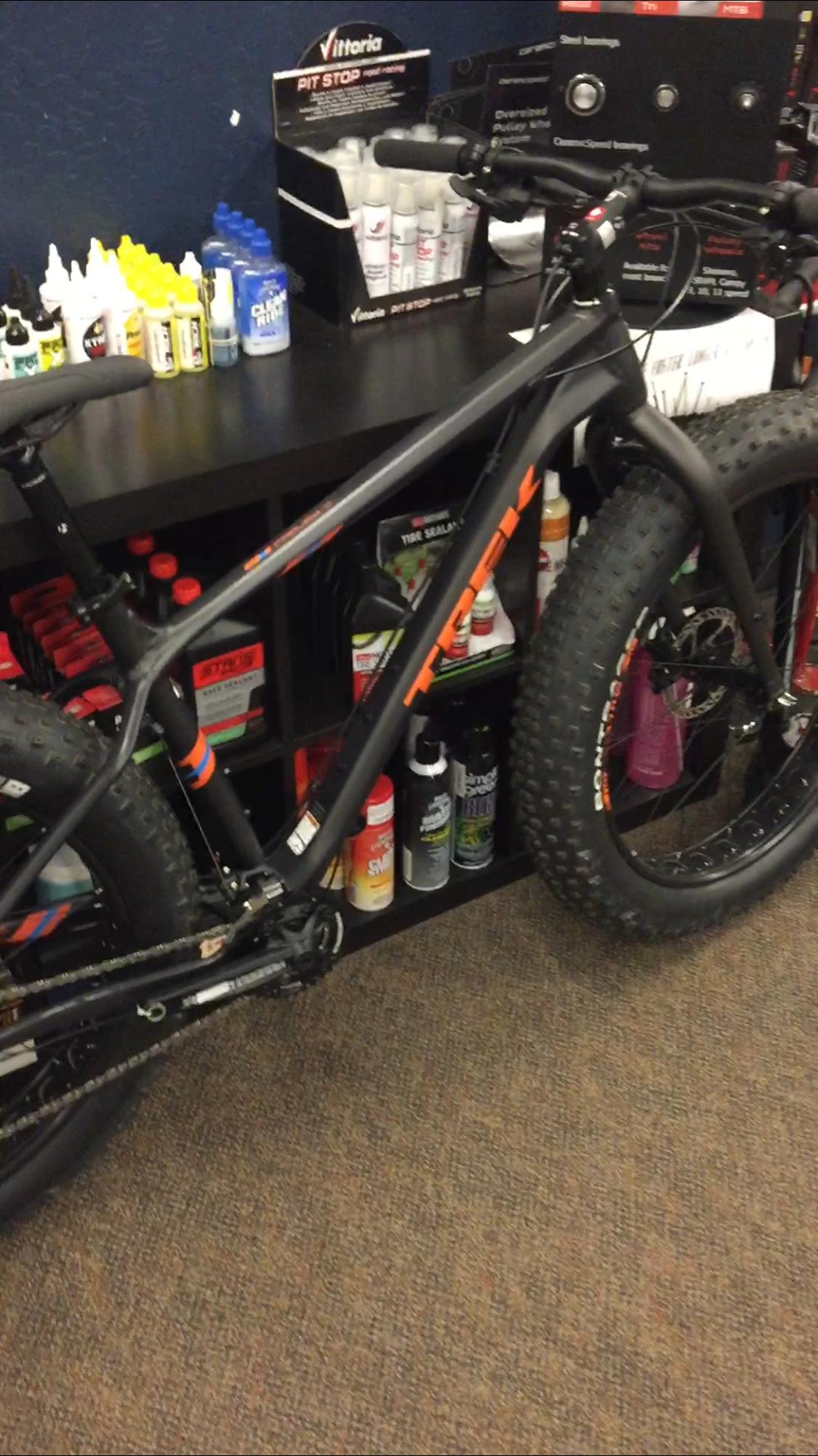 Trek farley 5: A black and orange fat bike is leaning against a shelf filled with various bicycle maintenance products, including lubricants and tire sealants. The background features a dark blue wall. The floor is covered in a textured carpet.