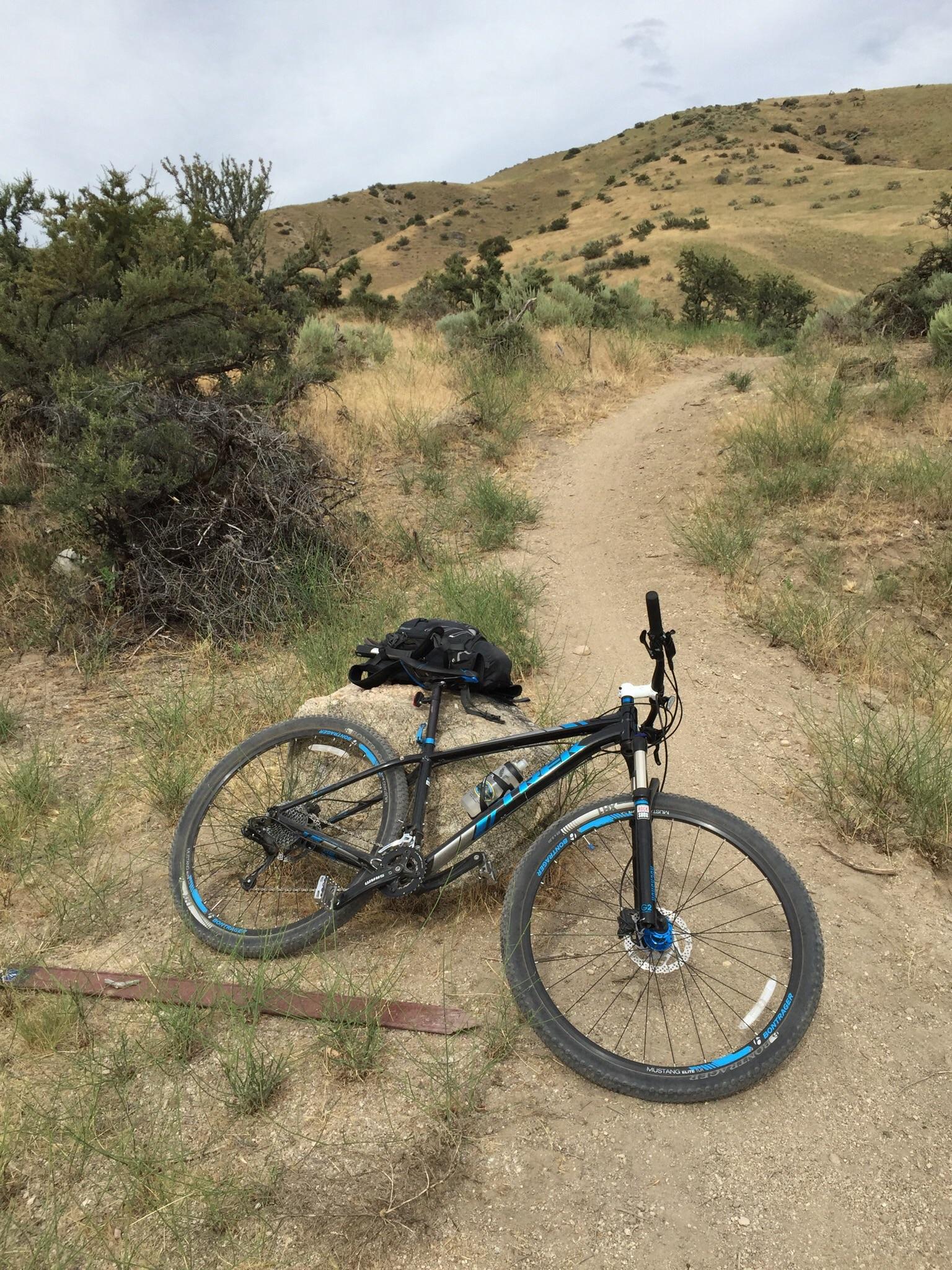 Trek Excalibur: A mountain bike resting on a dirt trail surrounded by tall grass and shrubs, with rolling hills in the background under a cloudy sky. A black backpack is placed nearby on the ground.