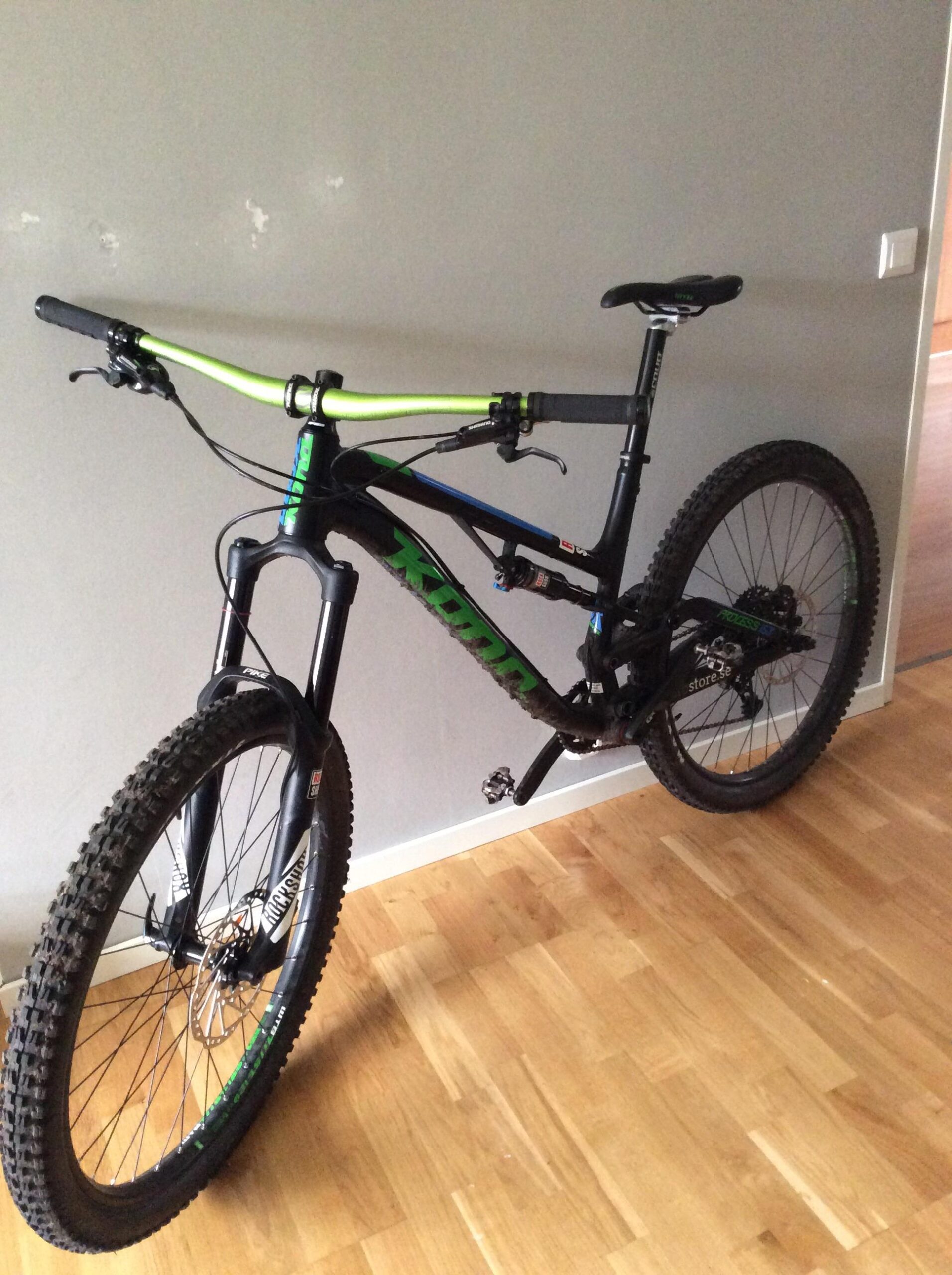 Kona Process 153: A black mountain bike with green and blue accents, leaning against a gray wall. The bike features thick tires and a front suspension fork, with handlebars that have bright green grips. The background shows a wooden floor and a partially visible interior space.