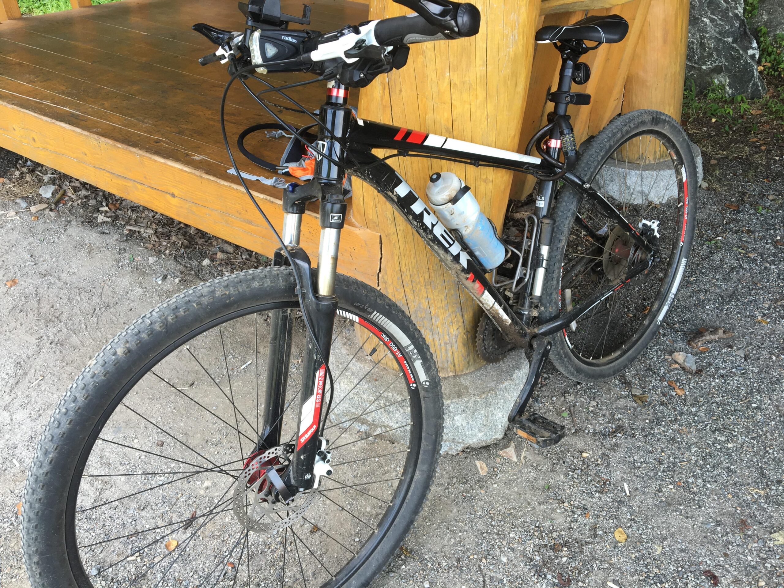 Trek Excalibur: A black and red mountain bike leaning against a wooden structure, with a water bottle attached to the frame. The bike has knobby tires and front suspension forks, sitting on gravel ground with patches of dirt.