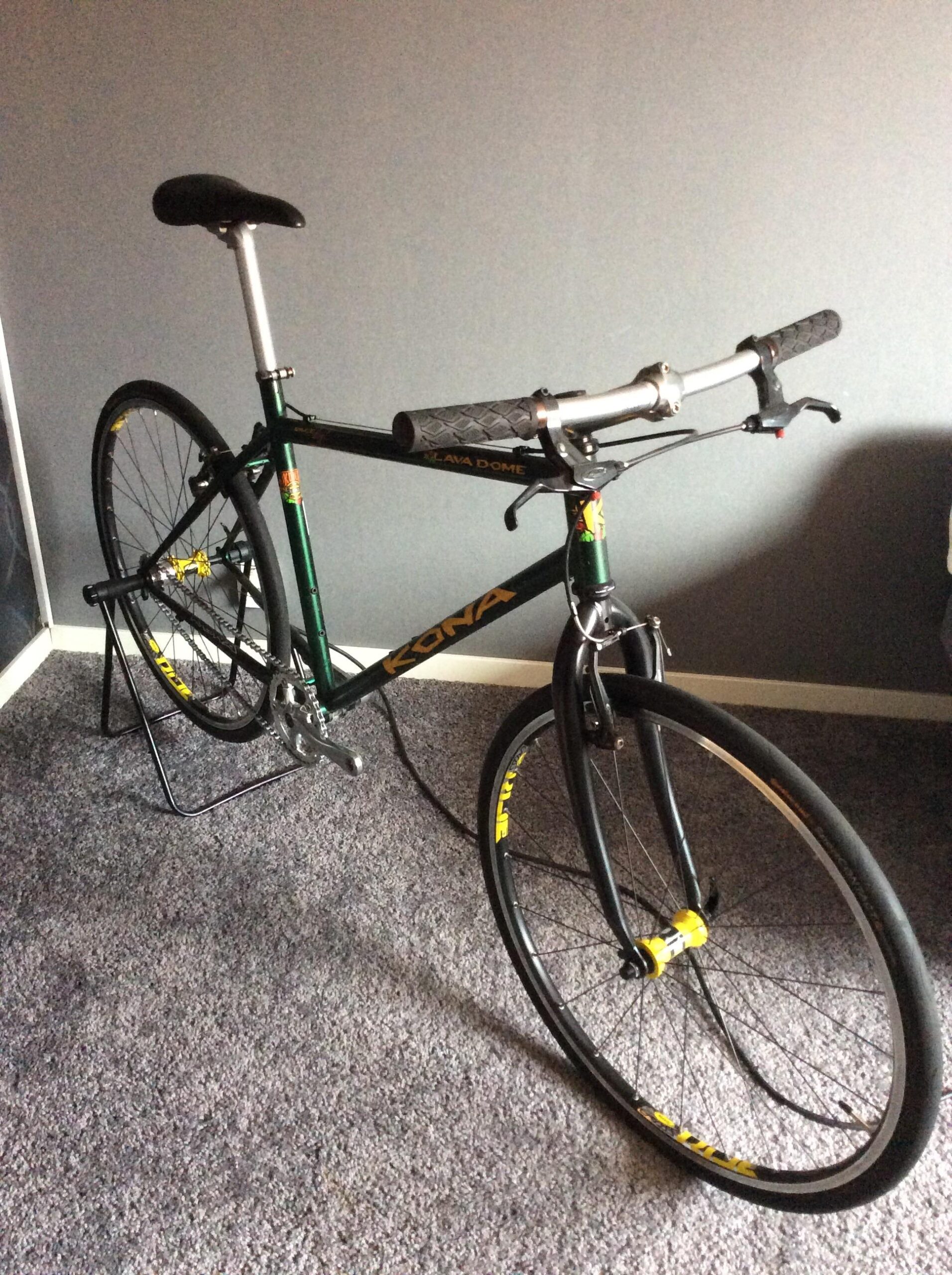 Kona Lava Dome: A green and black Kona Lava Dome bicycle is displayed on a stand against a gray wall. The bike features a minimalist design with sleek tires and a simple handlebar setup. It rests on a plush gray carpet.