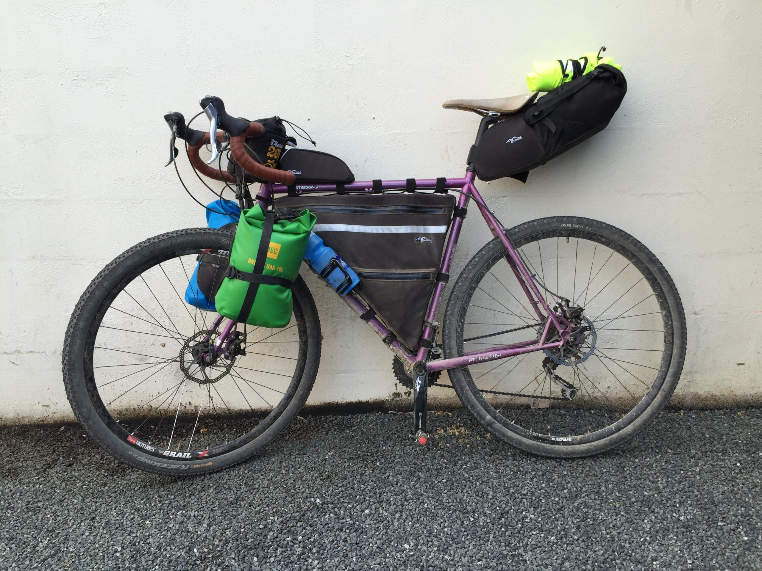 Surly Surly Straggler: A purple gravel bike with multiple bags attached, including a green dry bag on the frame, a black seat bag, and blue panniers on the handlebars, resting against a light-colored wall. The bike features knobby tires for off-road use and shows signs of gravel riding with dirt on the wheels and frame.