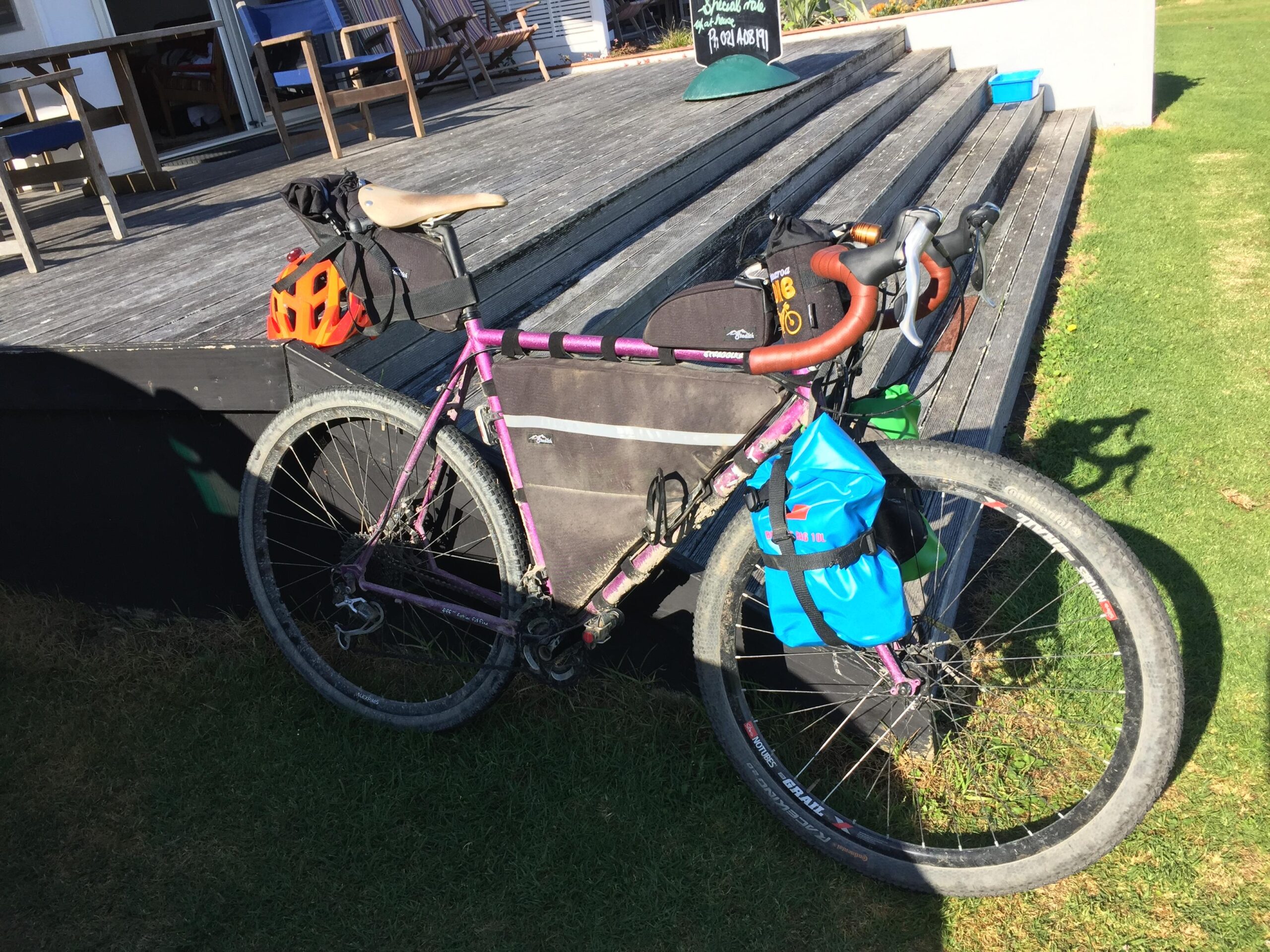 Surly Surly Straggler: A purple bicycle with a brown leather saddle is parked on grass near a wooden deck. It has various accessories attached, including a black bag on the frame, an orange helmet on the saddle, and a blue waterproof bag on the side. The scene is set outdoors on a sunny day, with wooden chairs visible in the background.