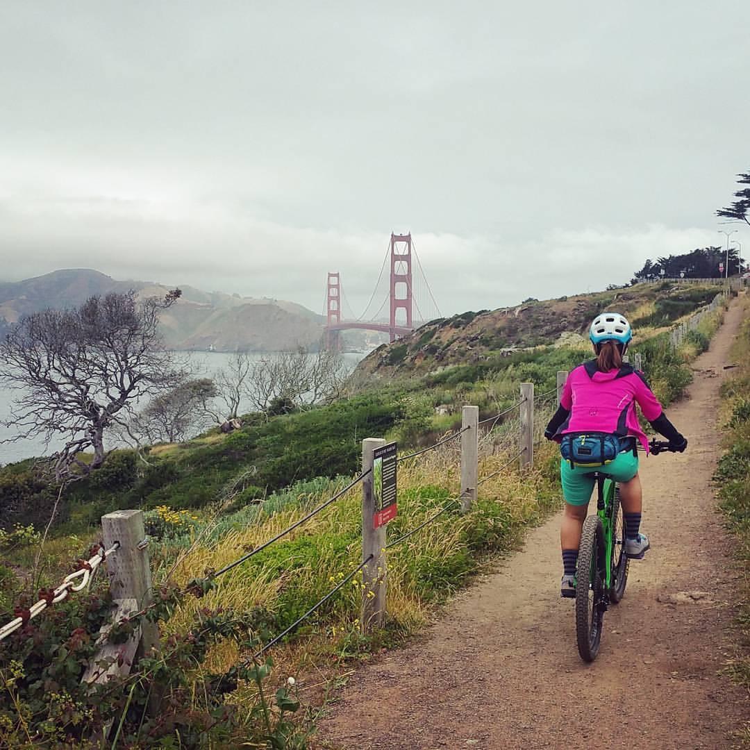 A cyclist wearing a pink jacket and helmet rides along a dirt path with greenery on either side, overlooking the Golden Gate Bridge in the distance on a cloudy day. Coast View Trail mountain bike trail.