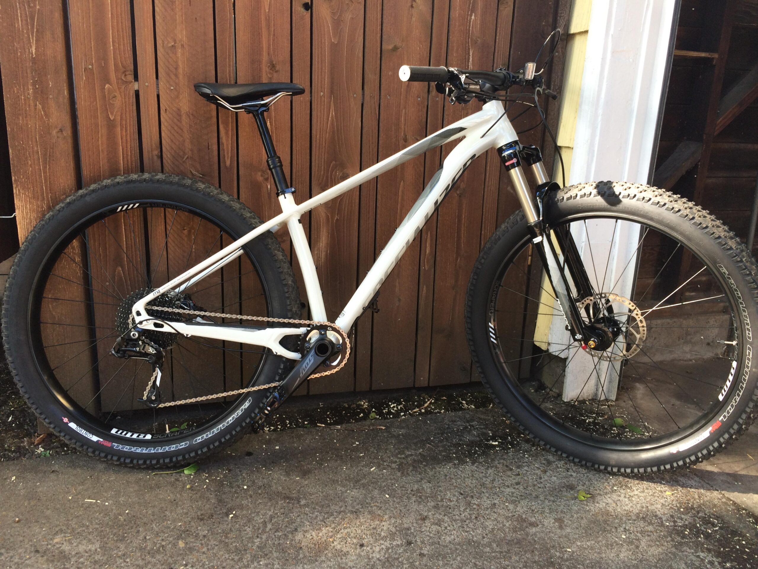 Specialized Fuse: A mountain bike with a white frame, black tires, and a visible suspension fork, positioned against a wooden fence. The bike features a single gear system and a black seat post, highlighting its design for off-road cycling.