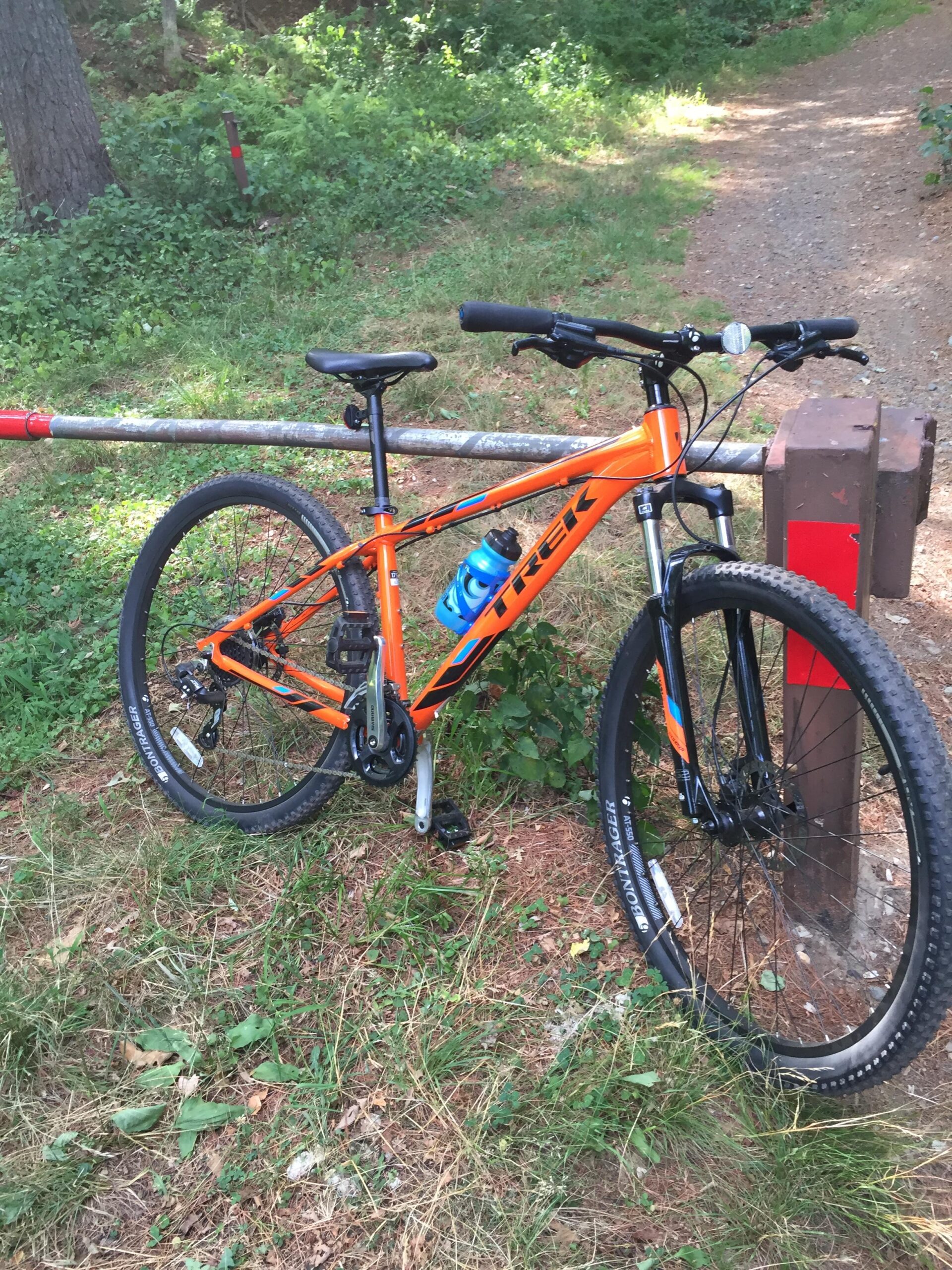 Trek Marlin 7: A bright orange mountain bike leaning against a wooden post in a grassy area surrounded by trees. The bike features black tires and a water bottle mounted on the frame. A dirt path is visible in the background, indicating a trail.