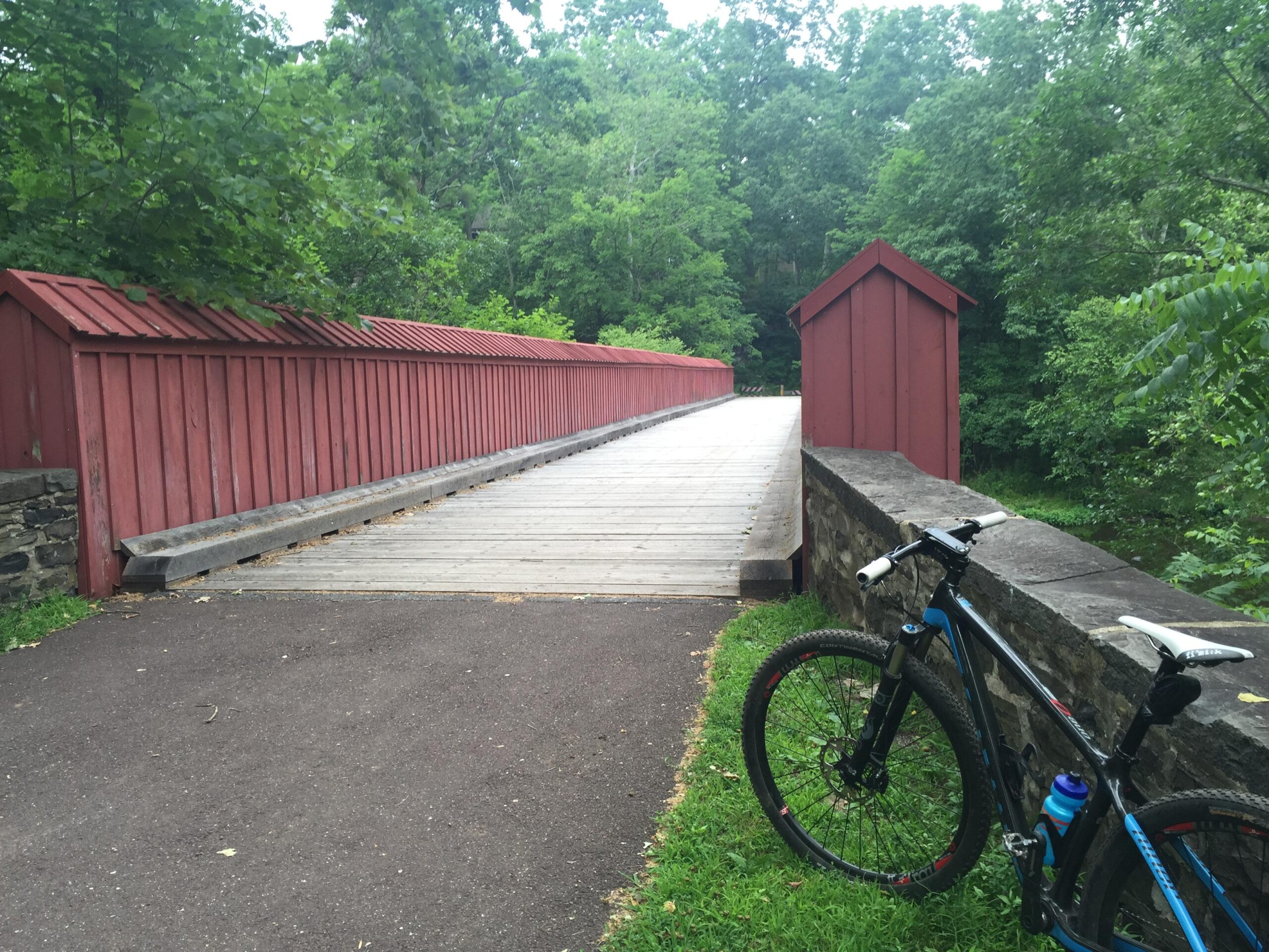 A red covered bridge spans a pathway surrounded by lush green trees. A mountain bike with a blue water bottle rests on the grass at the edge of the path, leading up to the bridge. High Rocks mountain bike trail.
