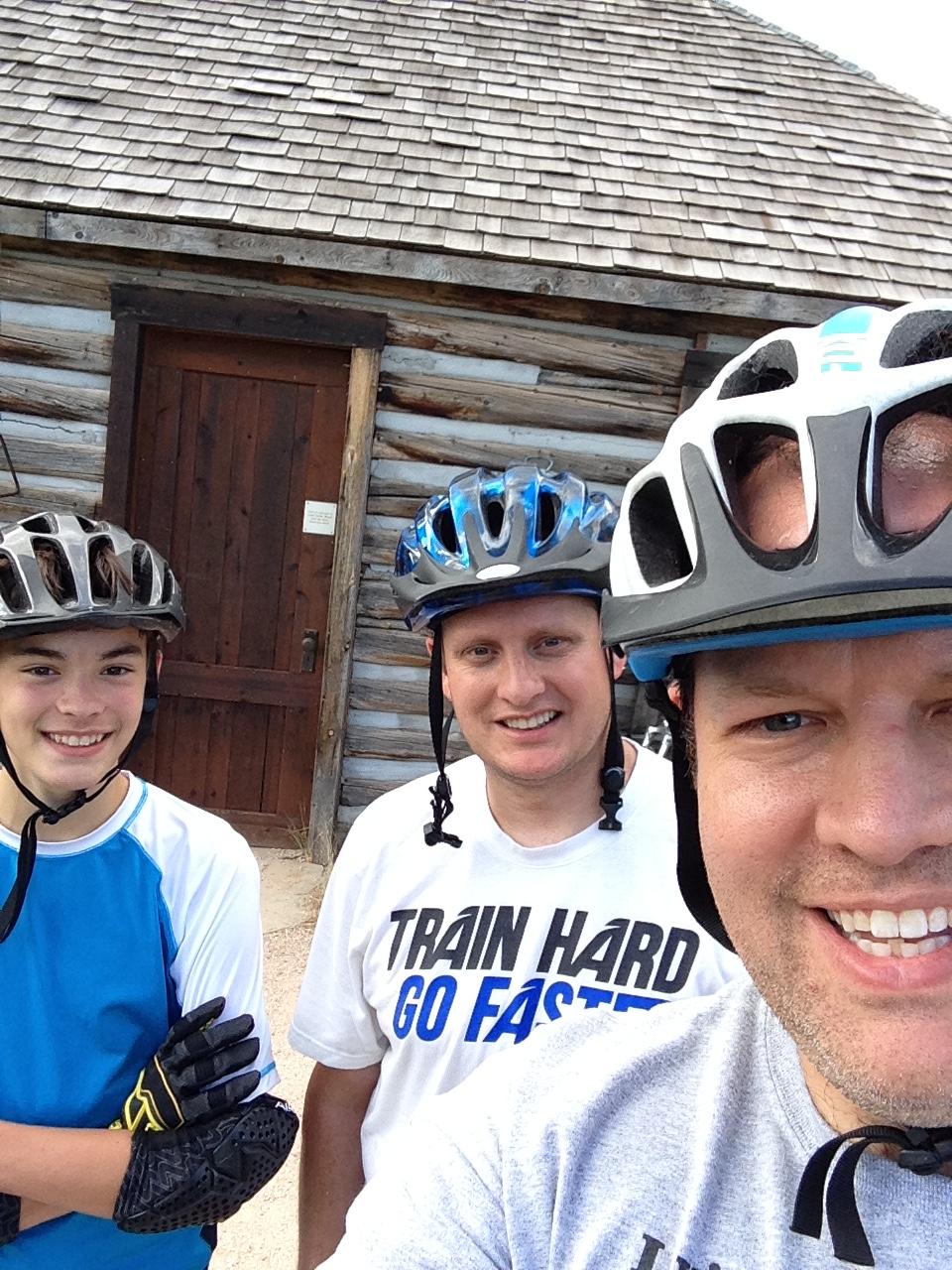 Three people wearing bike helmets pose for a selfie in front of a wooden building. Two individuals are smiling at the camera; one is an adult man in a gray shirt, and the other is a teenage boy in a blue shirt with black gloves. The third person, another adult man, is slightly behind and wearing a white shirt with the phrase "TRAIN HARD GO FASTER." The background features a rustic log building with a wooden door. Bobcat Ridge mountain bike trail.