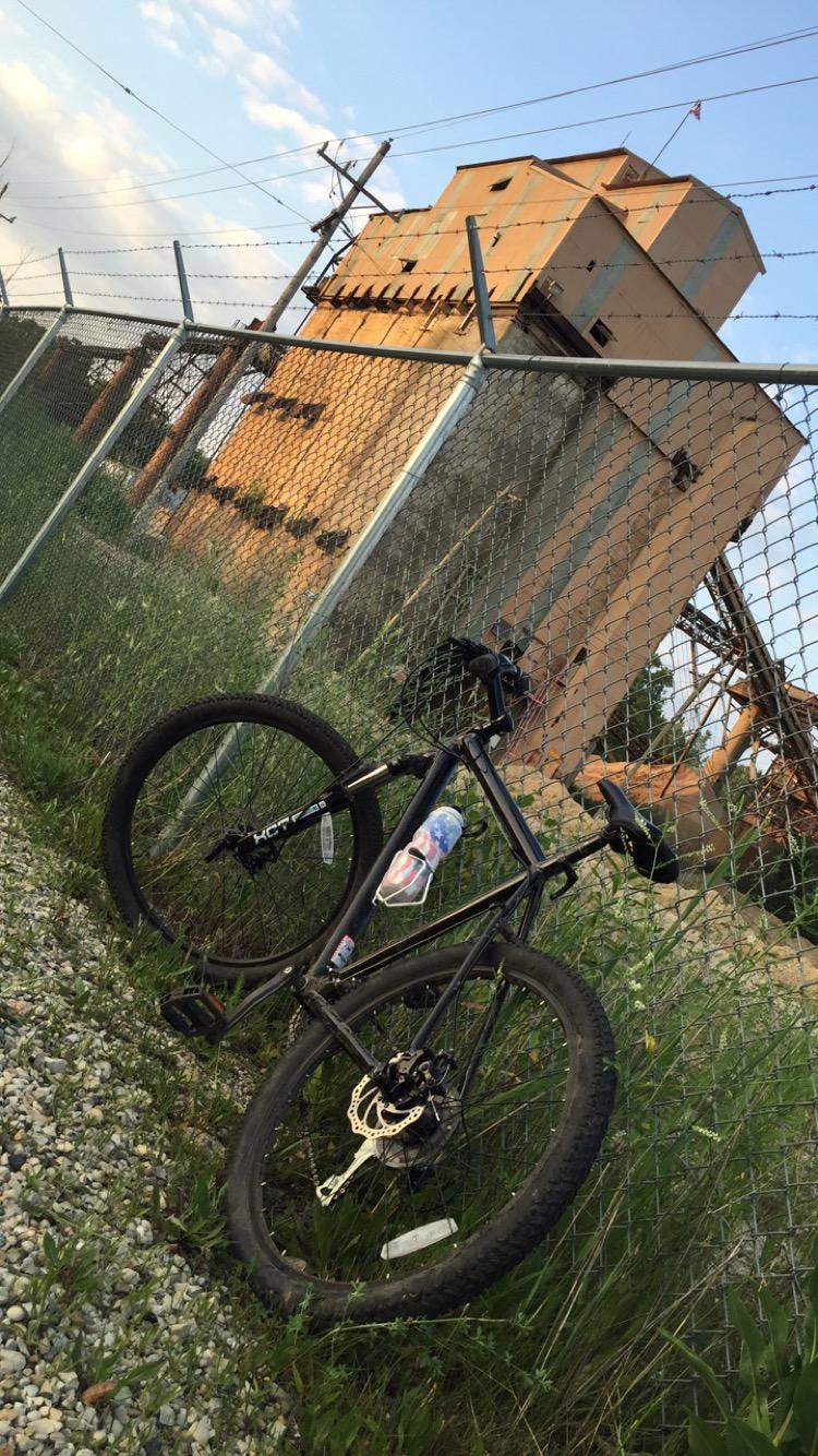 Nashbar AT-2: A black mountain bike is leaned against a chain-link fence, with an industrial building partially visible in the background. The building is large and rusty, surrounded by overgrown grass and gravel. The scene is set under a cloudy sky, suggesting late afternoon or early evening light.