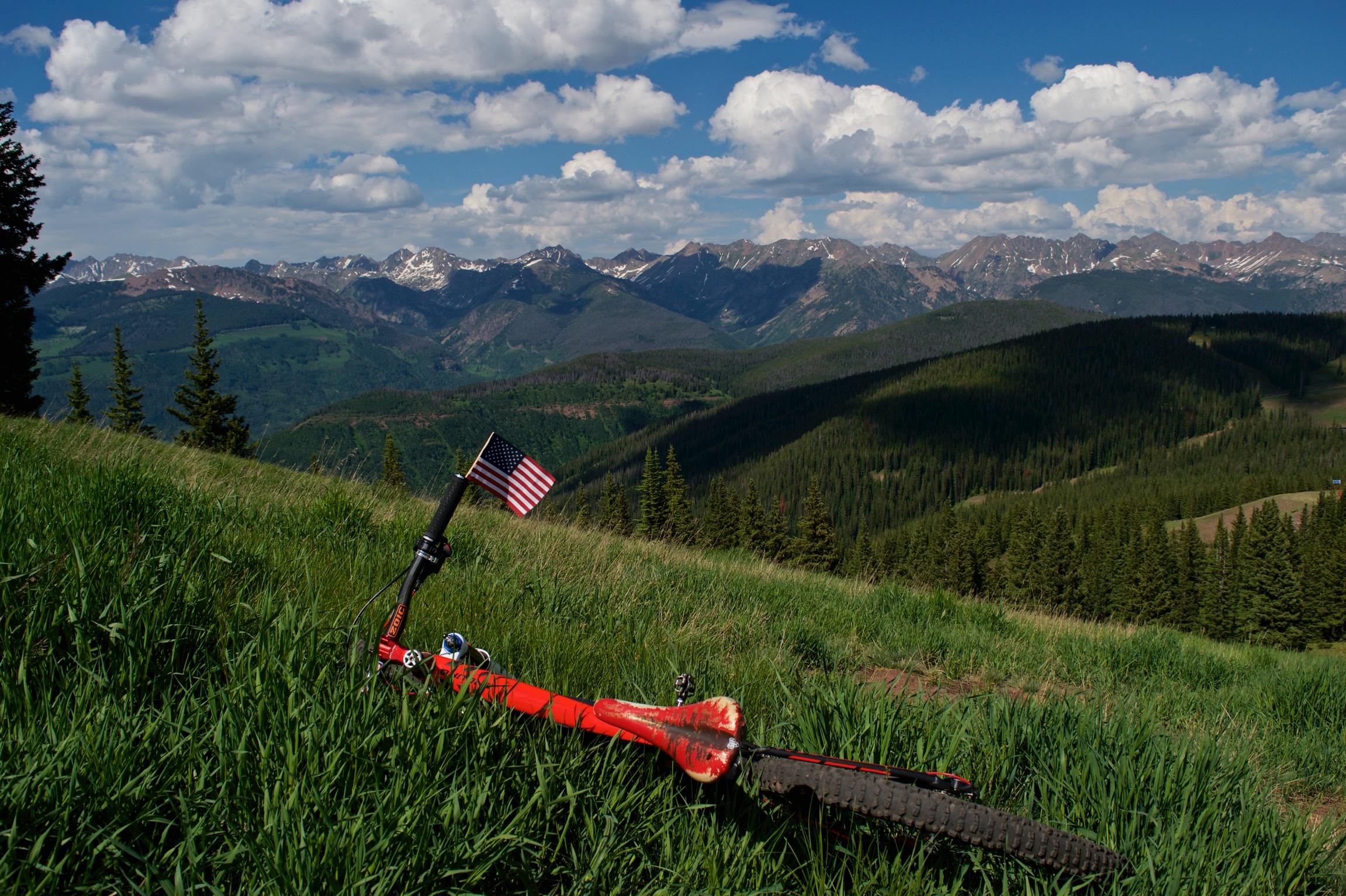 A vibrant red mountain bike rests on a grassy hillside, with an American flag attached to the handlebars. The background features expansive green mountains and a range of snow-capped peaks under a partly cloudy sky, showcasing a beautiful natural landscape. Vail Mountain Bike Park mountain bike trail.