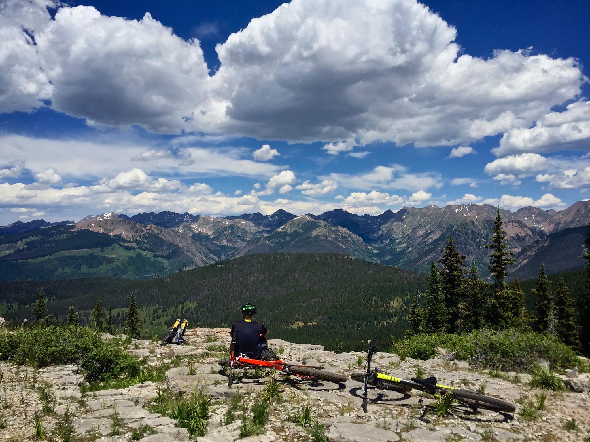 A person sitting on a rocky outcrop, overlooking a vast mountainous landscape with lush greenery and distant snow-capped peaks under a blue sky filled with fluffy clouds. Two mountain bikes are resting nearby. Vail Mountain Bike Park mountain bike trail.