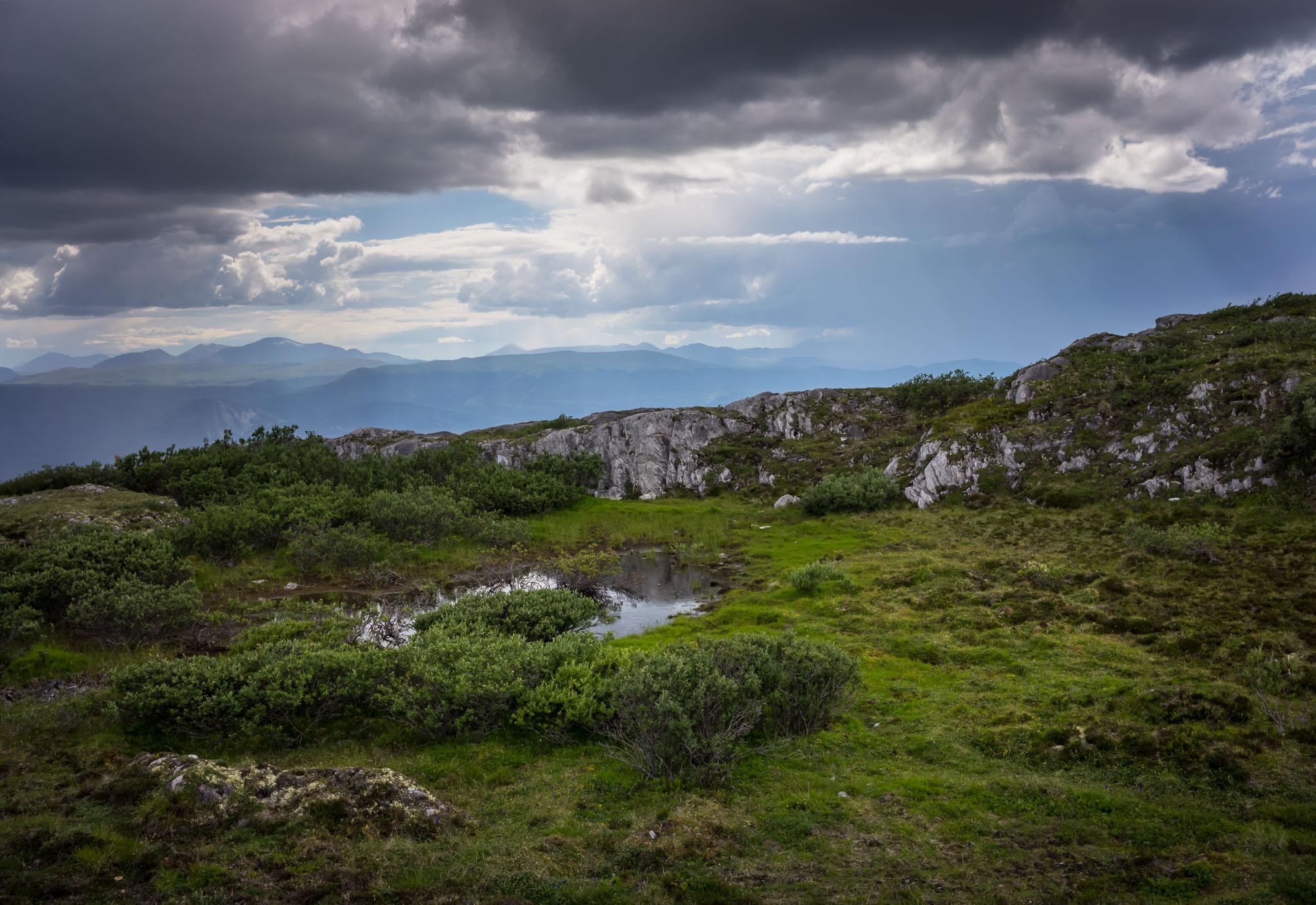 A scenic view of a mountainous landscape under a dramatic sky filled with clouds. In the foreground, lush green grass and shrubs surround a small pond, while rocky outcrops add texture to the terrain. In the background, distant mountains fade into the horizon. The atmospheric lighting creates a serene yet dynamic environment. Upper Boogaloo mountain bike trail.