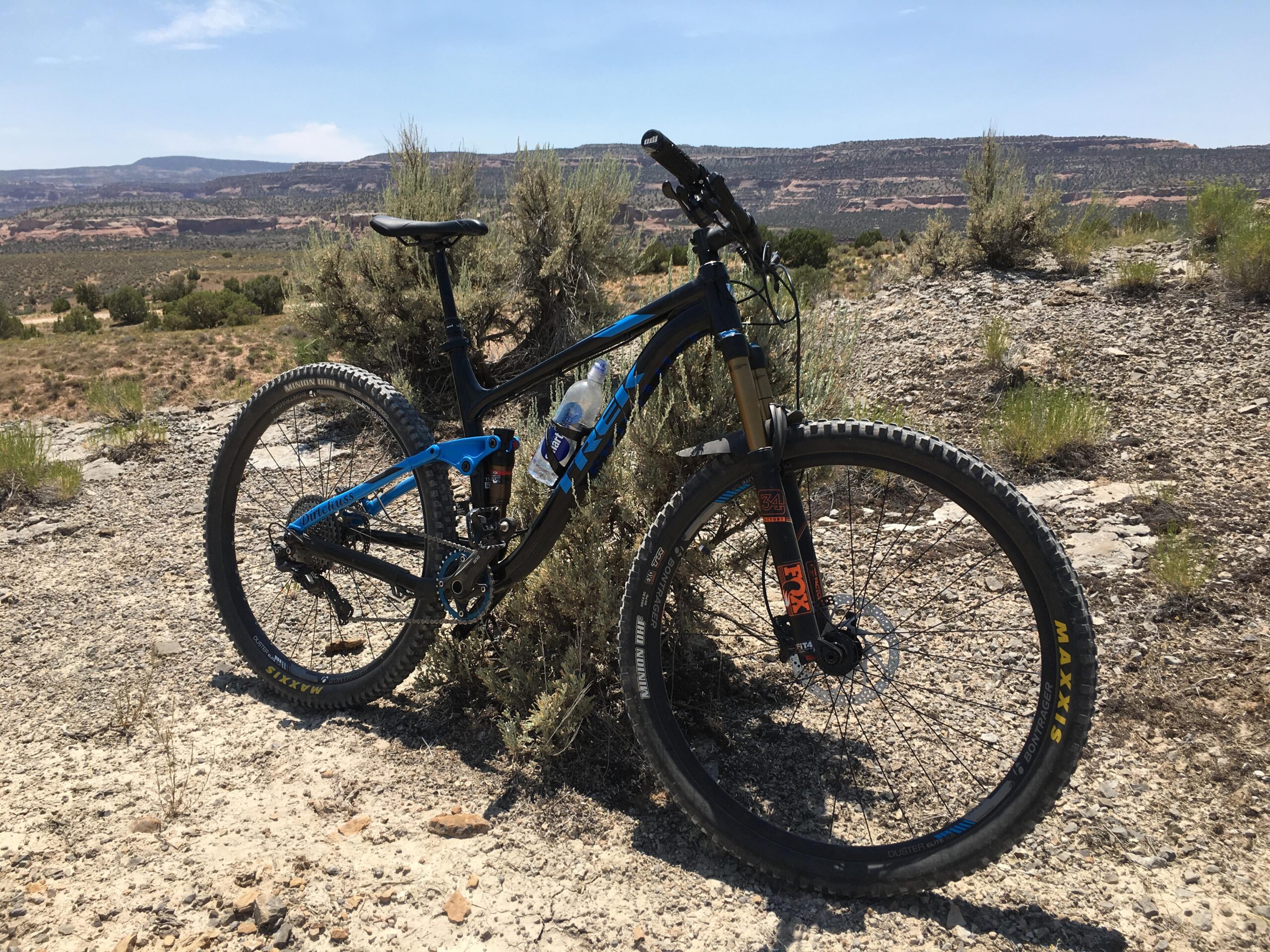 Trek Fuel EX 7 29: A mountain bike in a rocky, desert landscape, partially surrounded by shrubs and vegetation. The bike features a black and blue frame, front suspension, and wide tires, with a water bottle mounted on the frame. In the background, rolling hills and distant cliffs are visible under a clear blue sky.