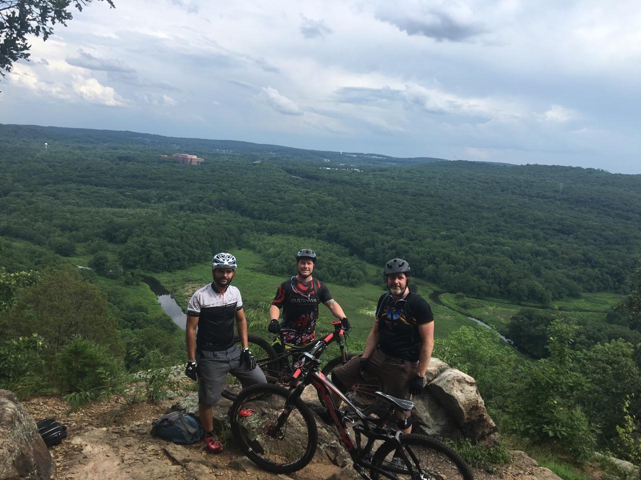 Three mountain bikers pose for a photo on a rocky outcrop overlooking a lush green valley and a river below. They are wearing helmets and biking gear, with a scenic view of trees and a distant hill in the background under a partly cloudy sky. Allamuchy State Park-North mountain bike trail.