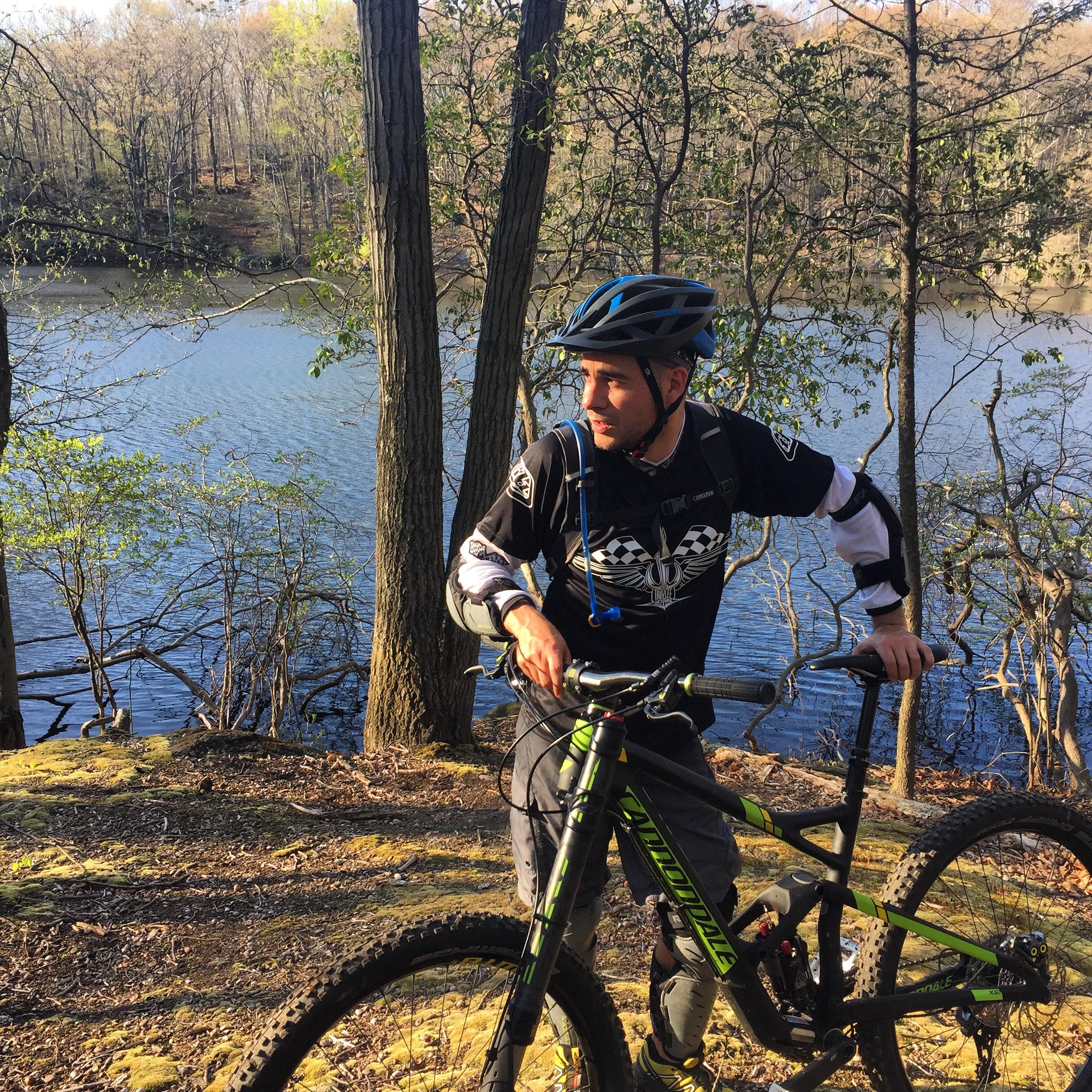 Cannondale Jekyll: A mountain biker in a helmet stands beside his bike near a calm lake, surrounded by trees. The scene captures the natural beauty of the outdoors with hints of early spring foliage.
