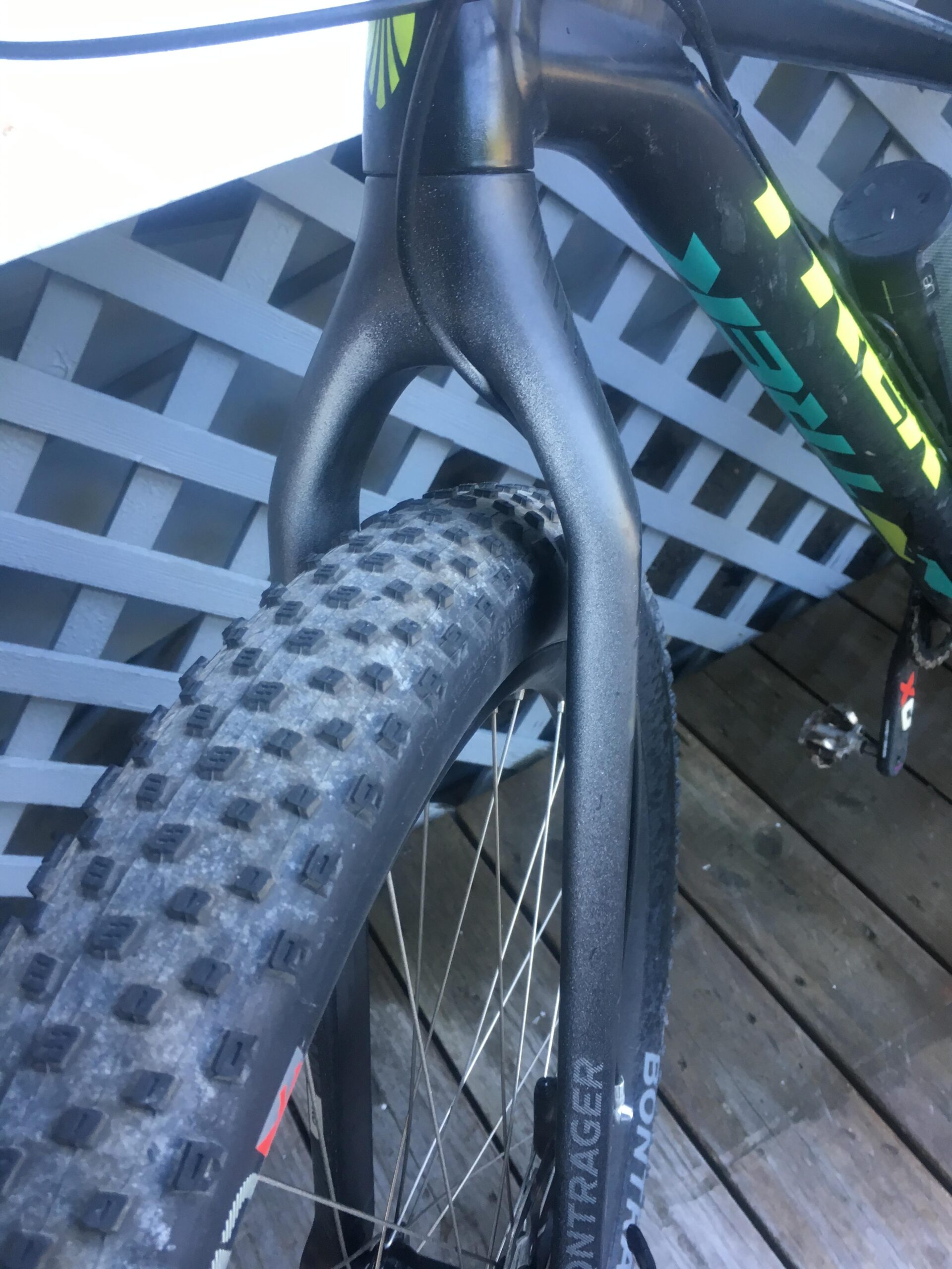 Trek Stache 9 29 Plus: Close-up of a mountain bike's front fork and tire, showcasing the sleek, black frame and textured tire tread. The background features a wooden deck and a lattice structure.
