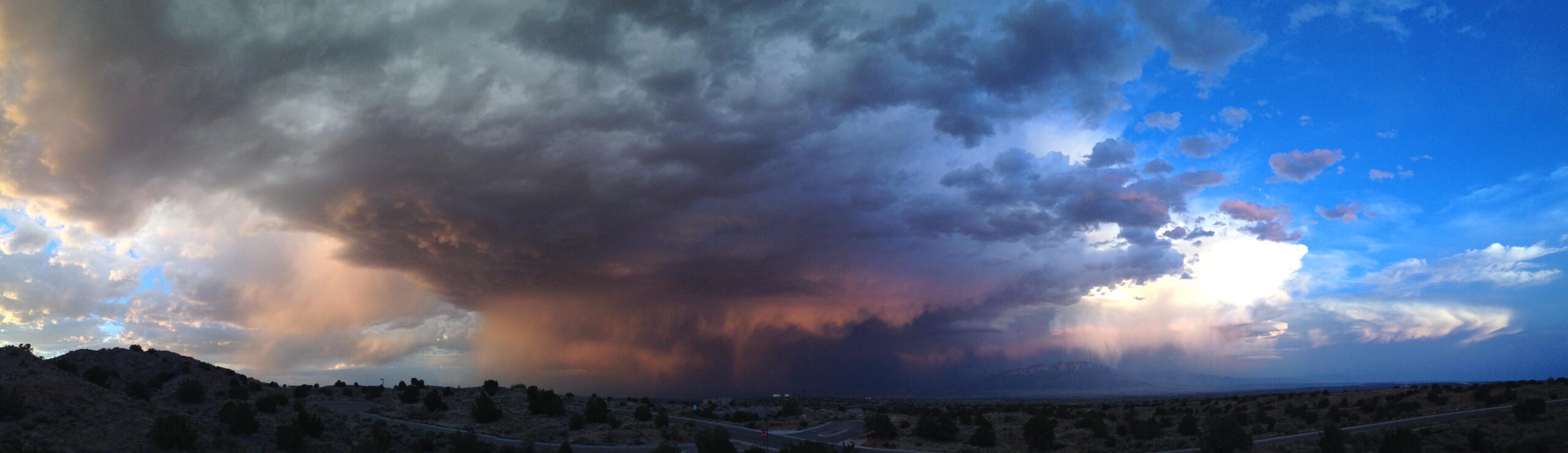 A panoramic view of a dramatic sky filled with dark storm clouds gradually transitioning to lighter shades and blue. In the distance, rain can be seen falling, creating a large curtain of precipitation against the backdrop of mountainous terrain. The scene captures a dynamic interplay of light and shadows, showcasing the beauty and intensity of an impending storm. Mariposa Fat Bike Trails mountain bike trail.