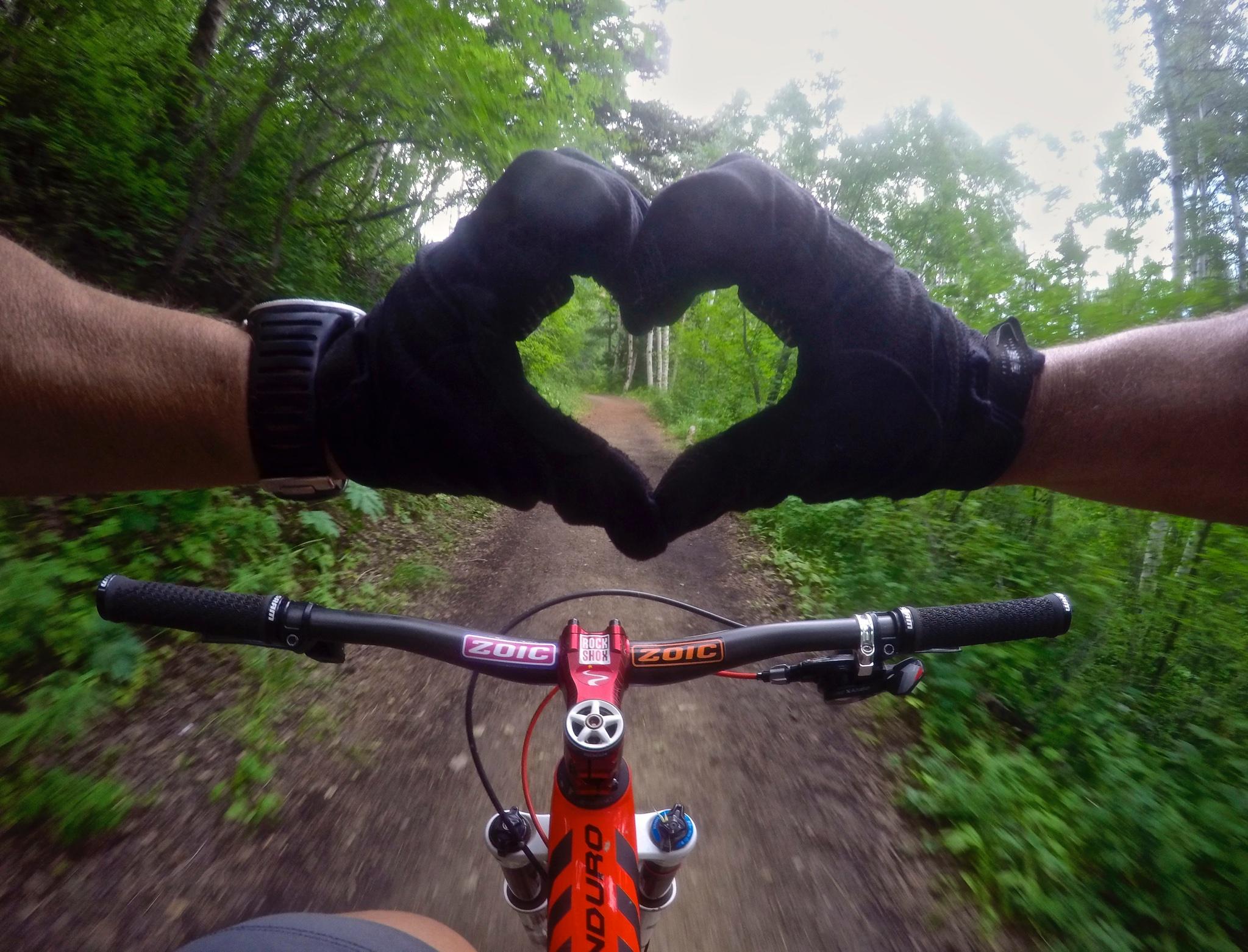 A close-up view of a mountain biker's hands forming a heart shape, with lush green foliage in the background. The cyclist is holding the handlebars of a bright red mountain bike, equipped with black grips and visible components, while riding along a dirt trail. Beaver Creek Ski Resort mountain bike trail.