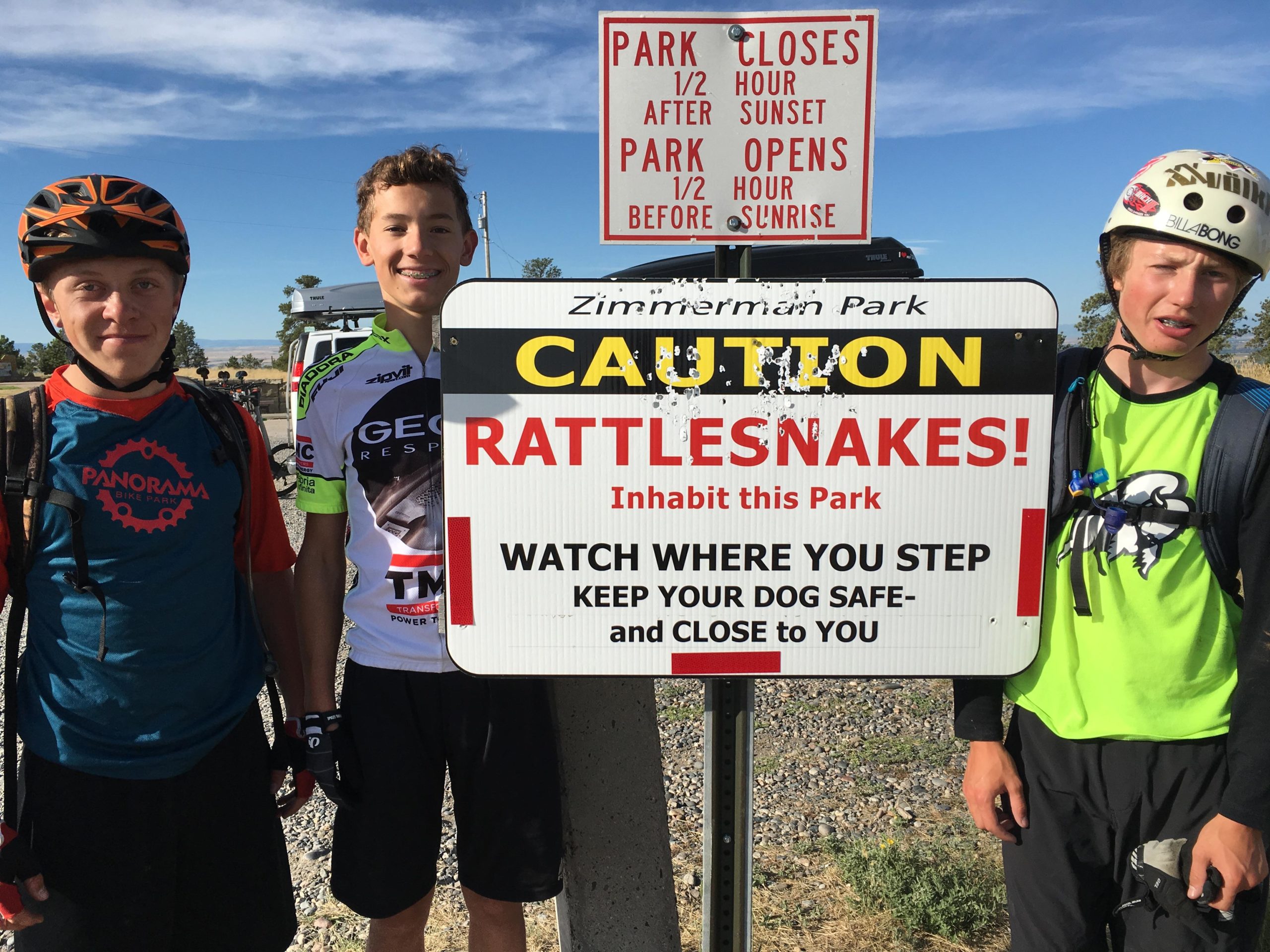 Three young boys in cycling gear stand next to a warning sign at Zimmerman Park. The sign reads "CAUTION RATTLESNAKES! Inhabit this Park" and includes additional safety instructions. The background features a clear blue sky and a gravel path leading into the park. Zimmerman Trail mountain bike trail.