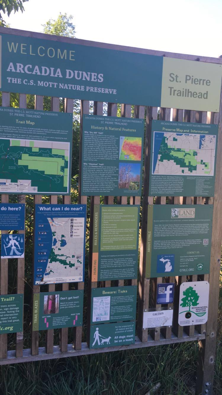 A wooden signboard at the St. Pierre Trailhead of the Arcadia Dunes C.S. Mott Nature Preserve, featuring information about the preserve, trail maps, rules and reminders for visitors, and tips for outdoor safety. The sign includes sections on history, natural features, and nearby activities, surrounded by lush greenery. Arcadia Dunes mountain bike trail.