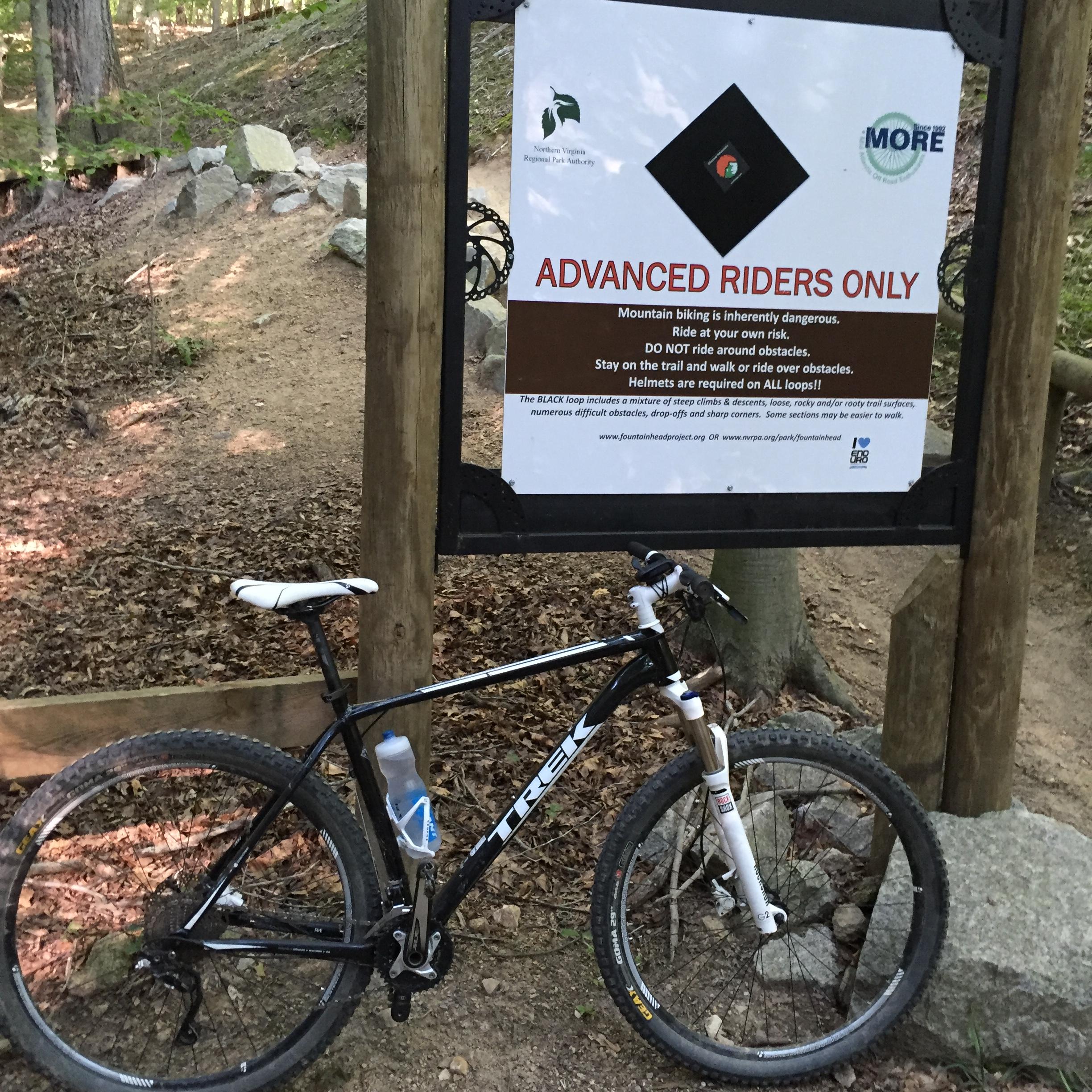 A mountain bike parked beside a "Advanced Riders Only" sign, which includes safety warnings and guidelines for mountain biking, set in a wooded area with a dirt trail and rocks in the background. Fountainhead Regional Park mountain bike trail.