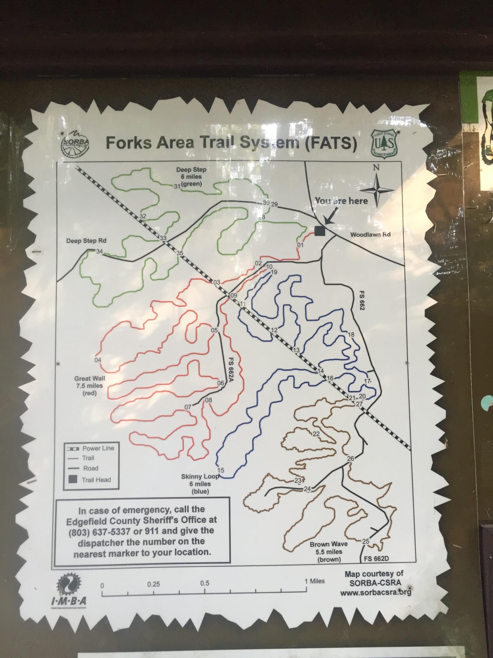Map of the Forks Area Trail System (FATS), displaying various marked trails in different colors: green for Deep Step, red for Great Wall, blue for Skinny Loop, and brown for Brown Wave. The map includes a key for trail types and emergency contact information. An indication of the viewer's current location is marked as "You are here" near Woodlawn Road. Forks Area Trail System (FATS) mountain bike trail.