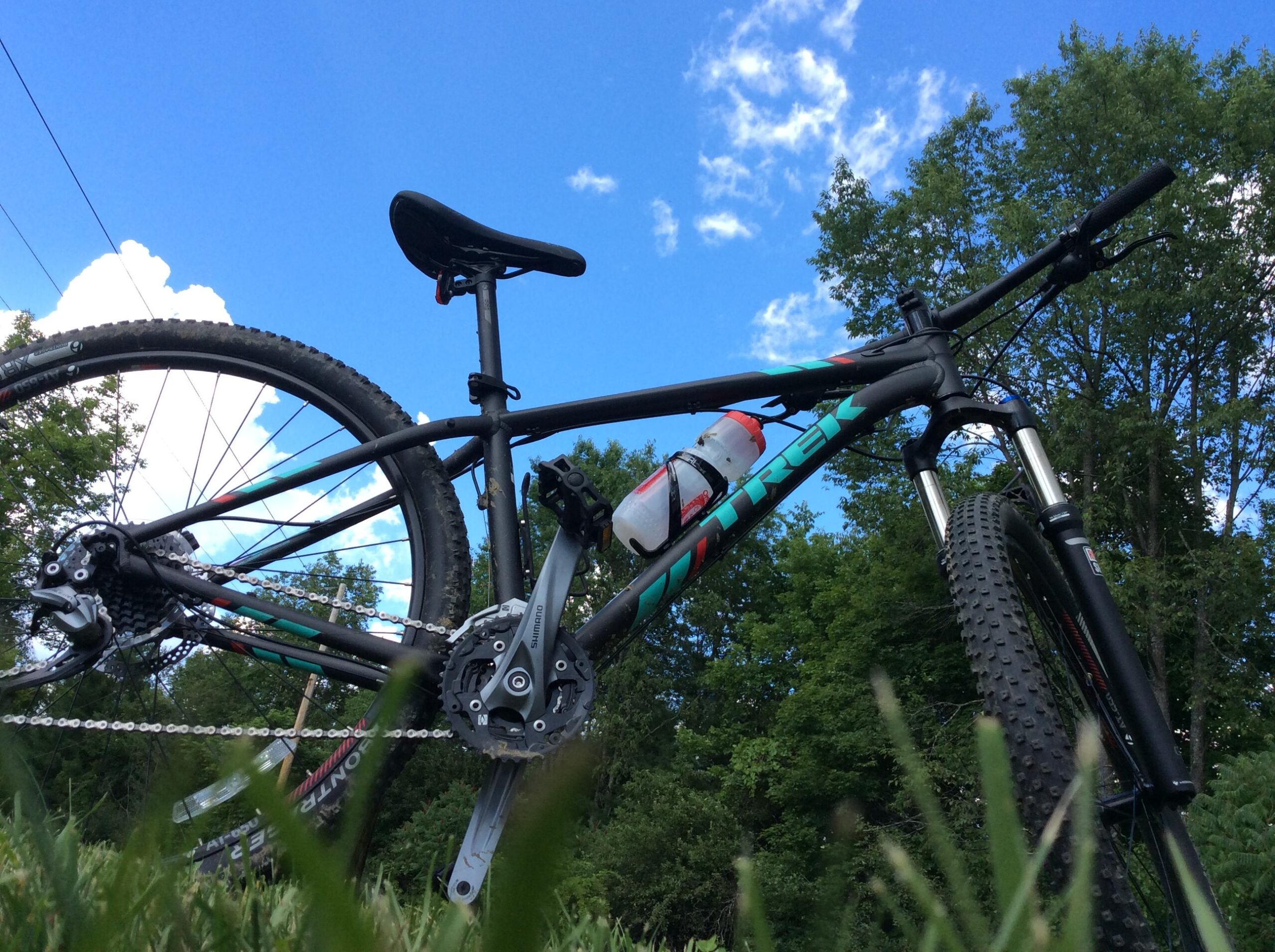Trek X-Caliber 7: A close-up view of a black mountain bike resting on grass, featuring a prominent water bottle attached to the frame. The background includes a bright blue sky with white clouds and green trees. The bike is positioned at an angle, showcasing its wheels and drivetrain components.