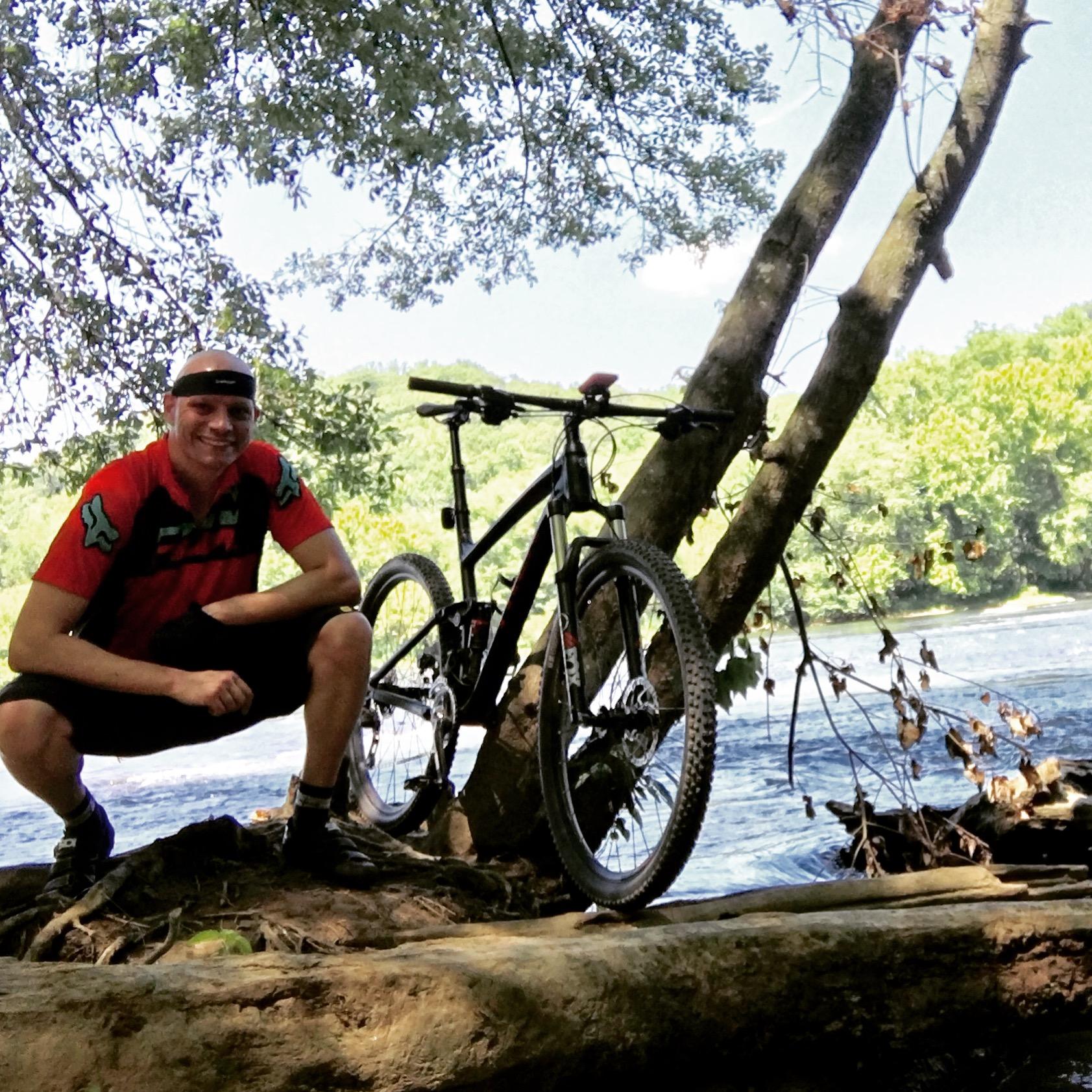 A person squatting next to a black mountain bike, posed on a log by a river. They are wearing a red and black athletic shirt and shorts, with trees and a lush green landscape in the background. Bright sunlight filters through the leaves above. Sope Creek mountain bike trail.