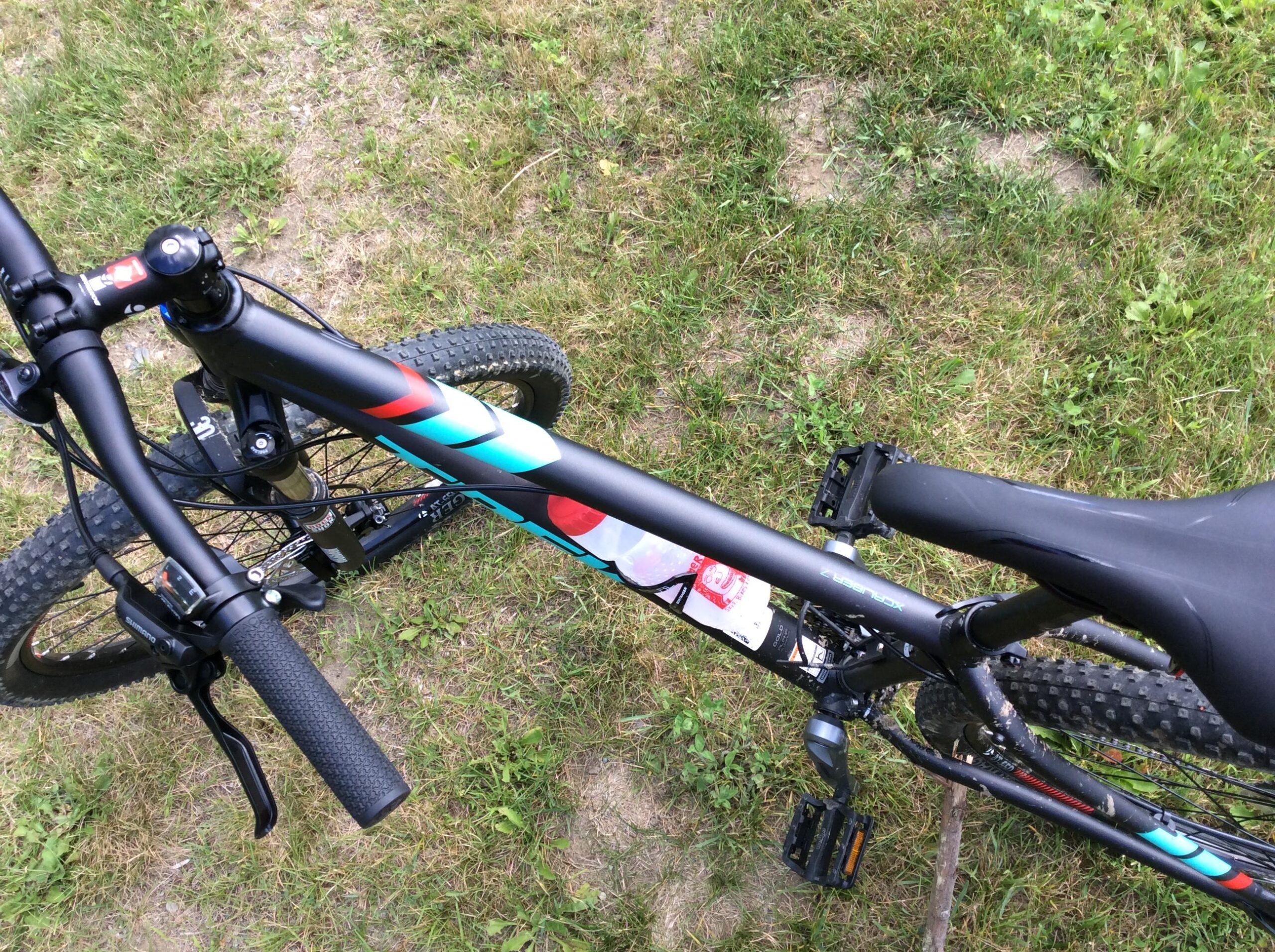 Trek X-Caliber 7: A close-up view of a mountain bike positioned on grassy terrain. The image captures the handlebars, part of the frame with colorful graphics, and the seat. The bike features black tires with prominent treads and a water bottle attached to the frame.