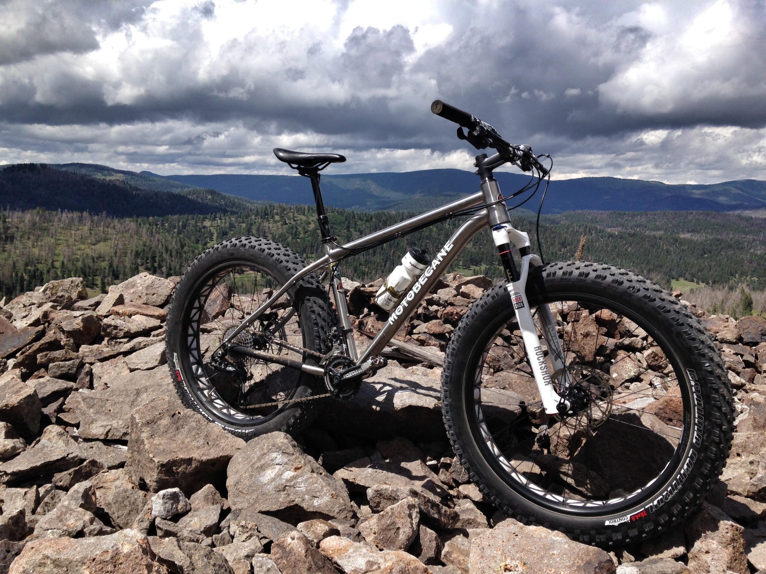 A mountain bike with a sturdy frame and wide tires is positioned on a rocky outcrop, overlooking a lush green valley and distant mountains under a partly cloudy sky. Valles Caldera Trails mountain bike trail.